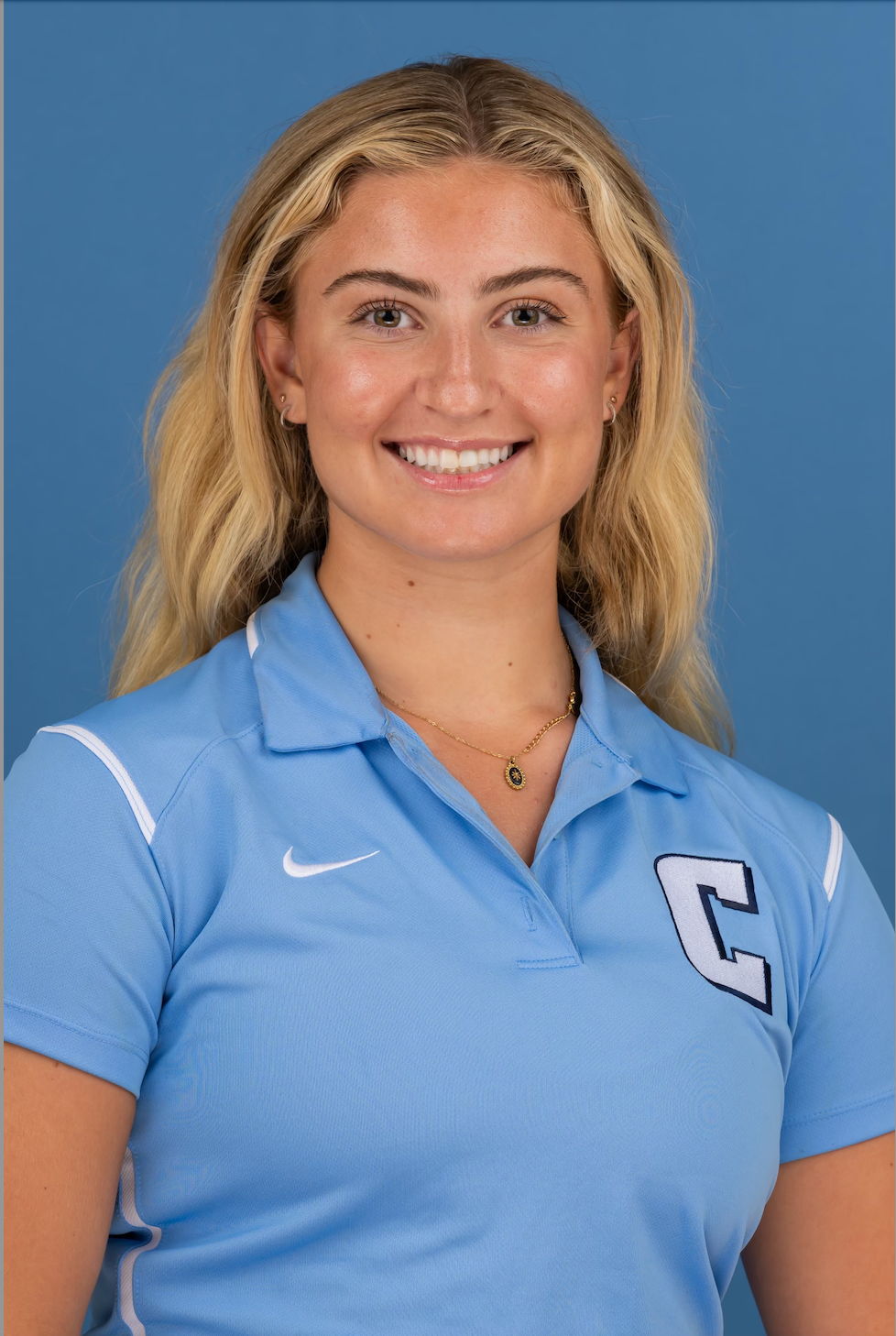 The image features a young woman with long, wavy blonde hair, smiling confidently at the camera. She is wearing a light blue polo shirt adorned with a prominent 'C' and the Nike logo, suggesting she may be associated with a sports team or school, possibly a captain given the letter's significance. The background is a solid blue, complementing her attire and enhancing the cheerful, approachable vibe of her demeanor. Her expression conveys warmth and enthusiasm, making her appear welcoming and friendly. 

The simplicity of the background highlights her as the main focus, indicating this could be a promotional or team photo. The overall composition suggests a context related to sports or school spirit, resonating with themes of teamwork and leadership. It's like she's ready to tackle any challenge—just don’t ask her about her favorite pizza topping; that’s a team divide waiting to happen!