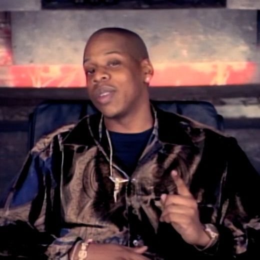 The image captures a young Jay-Z, exuding a confident and slightly mischievous demeanor. He's caught mid-gesture, pointing his right index finger as if emphasizing a crucial point, perhaps a lyrical gem or a business strategy. His expression is a subtle smirk, hinting at self-assuredness. He wears a distinctive patterned shirt with dark blue and brown hues, a dark undershirt, and a prominent gold chain with a unique pendant. A gold bracelet and watch adorn his left wrist. The background is a blurred, gritty urban wall, possibly concrete, with streaks of red light or graffiti, creating an edgy, industrial vibe typical of early hip-hop music videos. This setting perfectly complements his street-smart persona. The humor lies in his intense, almost professorial pointing, making one wonder if he's about to drop a groundbreaking verse or just remind someone to pick up milk.