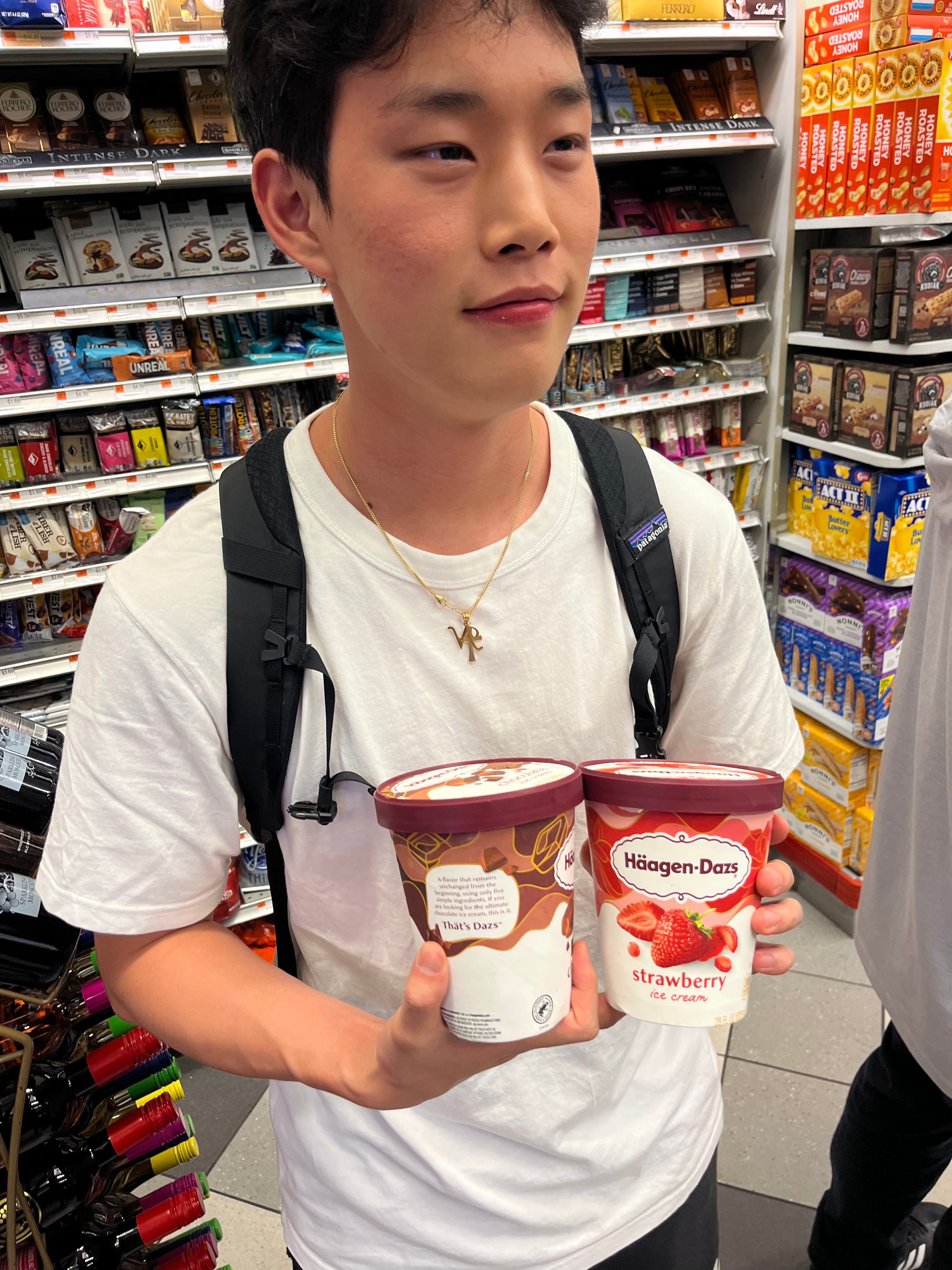 A young man, wearing a white t-shirt and a Patagonia backpack, stands in a brightly lit grocery store aisle, holding two large tubs of Häagen-Dazs ice cream: one chocolate and one strawberry. His demeanor is contemplative, with a slight upward gaze, as if pondering the profound implications of his sweet selections. The background is a vibrant display of popular brands like Ferrero Rocher, Ghirardelli, Honey Bunches of Oats, and Act II, perfectly setting the scene for a serious dessert decision. He appears to be on a singular mission for frozen indulgence, humorously embodying the intense focus one reserves for life's most delicious choices.