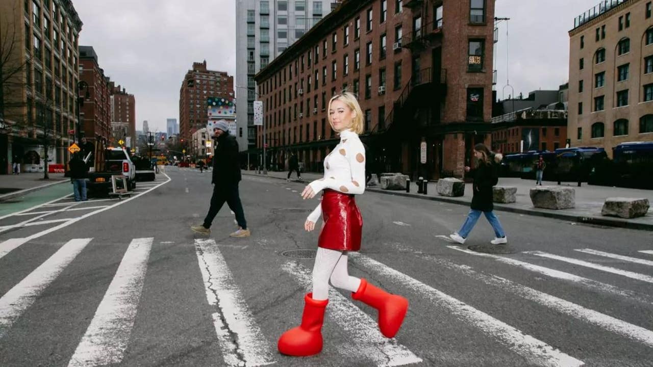 In the image, a young woman confidently walks across a city street, sporting a playful outfit that features a white top adorned with polka dots and a shiny red skirt. Her oversized, bright red footwear adds a whimsical touch, almost resembling cartoonish clown shoes, which contrasts with the more muted urban backdrop.
The surrounding architecture showcases a blend of modern and historic buildings, typical of a vibrant city like New York. The sky is overcast, hinting at a chilly day, yet her demeanor is cheerful, as she appears to embrace the moment with a light smile.
Pedestrians in the background add to the scene's bustling atmosphere, while a parked truck and traffic signals hint at the everyday life of the city. This playful juxtaposition of style and setting creates a humorous effect, as her bold fashion choice stands out against the more subdued urban environment.