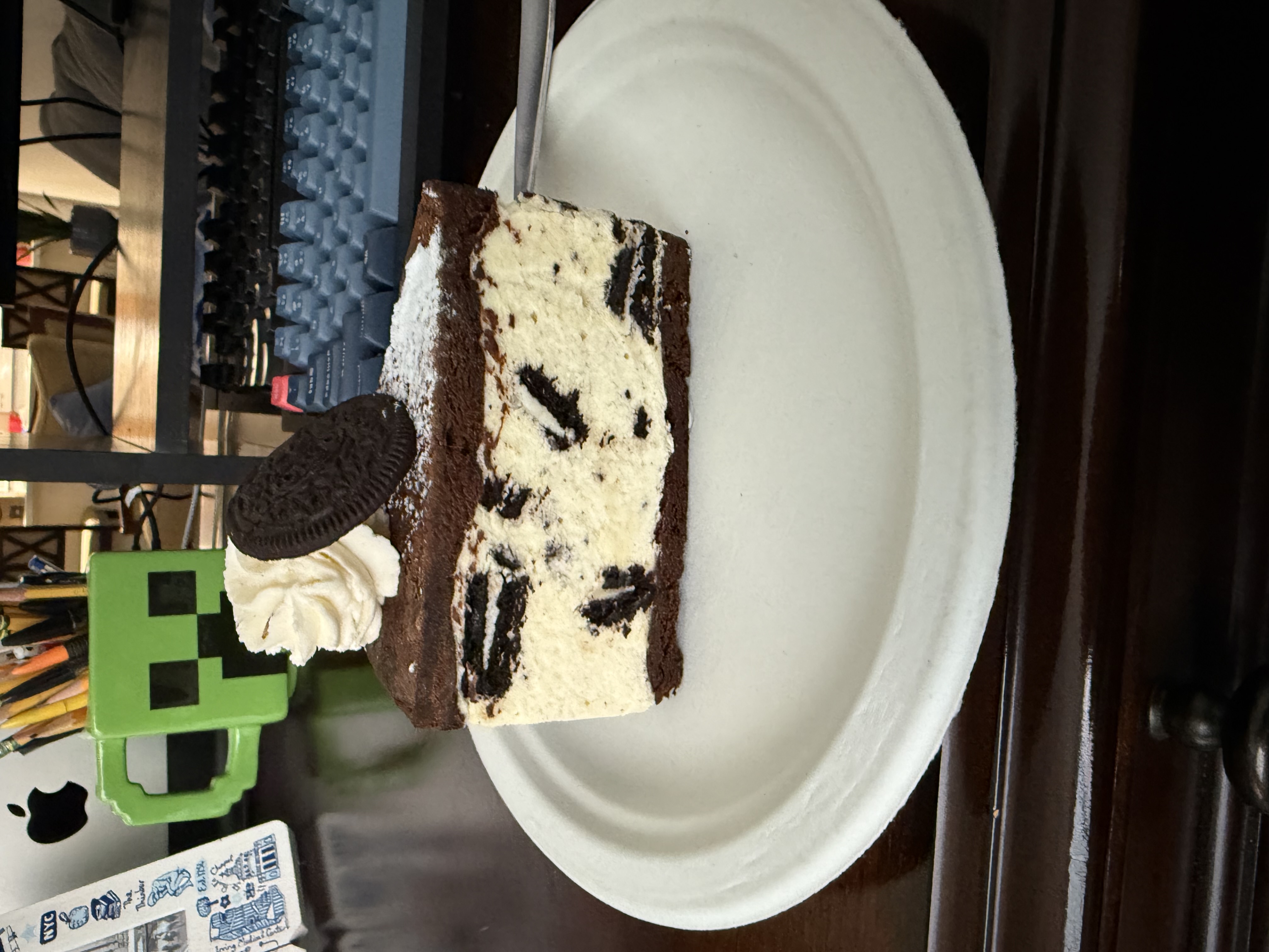 The image showcases a delightful slice of Oreo cheesecake, presented on a white paper plate. The cake features a rich chocolate crust topped with creamy layers infused with chunks of Oreo cookies, giving it an indulgent texture. A dollop of whipped cream and an Oreo cookie garnish sits atop the cake, adding a playful touch.
In the background, a green Minecraft Creeper mug stands out, hinting at a fun, gaming-inspired environment. This adds a whimsical element, suggesting that the setting is likely a casual workspace or a cozy home. The presence of a mechanical keyboard and a glimpse of a desk filled with various items further emphasizes this laid-back atmosphere.
Overall, the scene conveys a delightful blend of indulgence and creativity, inviting viewers to enjoy both a tasty treat and a playful gaming vibe—perfect for any Oreo fan or gamer!