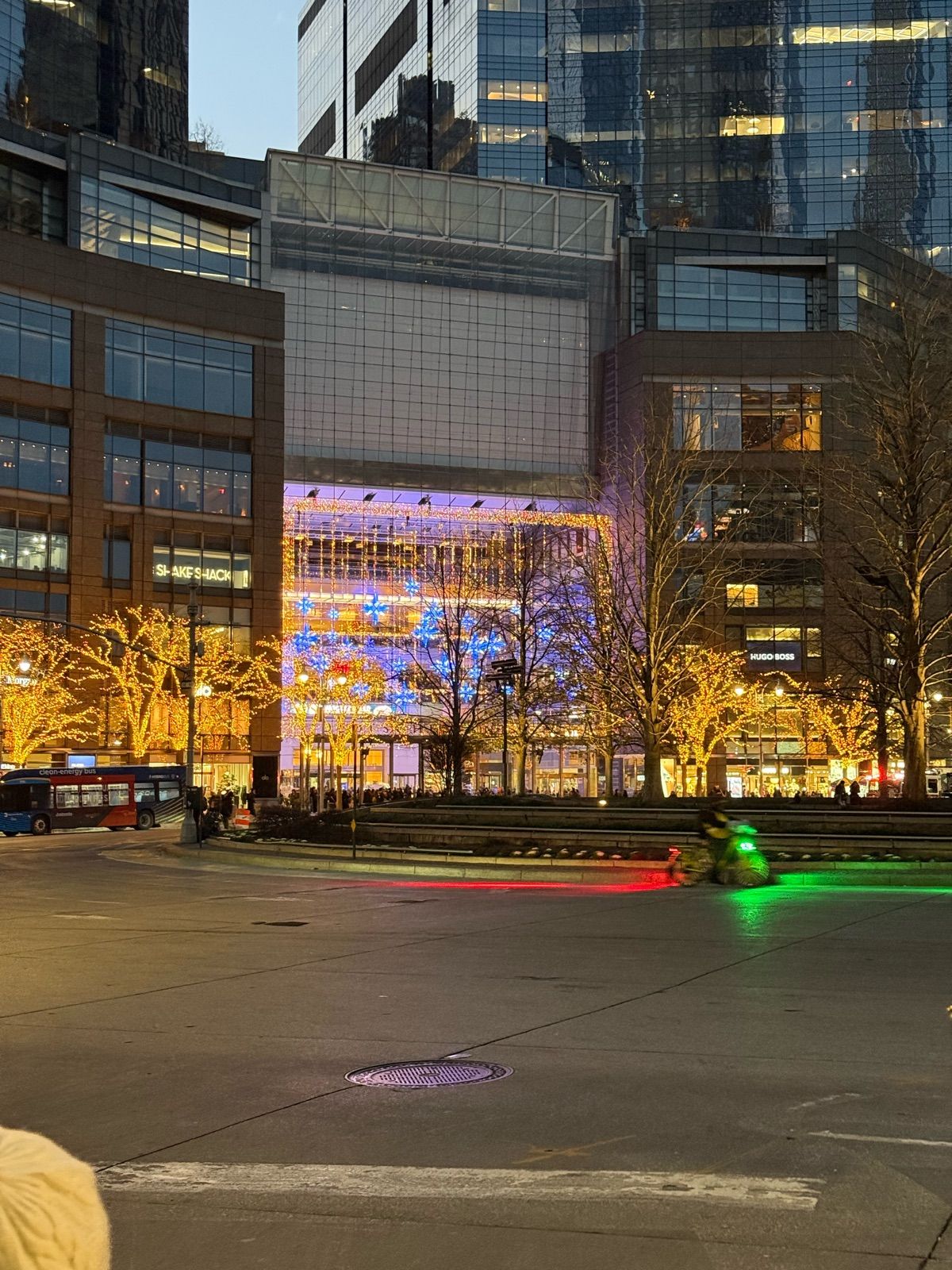The image captures a bustling urban scene at dusk, highlighting a modern building facade adorned with colorful lights. The main structure is illuminated with vibrant hues of blue and purple, suggesting a festive or celebratory atmosphere, possibly related to a holiday or special event. The surrounding trees are also embellished with warm, twinkling lights, enhancing the lively ambiance.
In the foreground, a bus waits at a stop, while a cyclist zooms by, leaving a trail of red and green light—adding a dynamic element to the composition. The people visible near the bus hint at a busy city life, perhaps gathering for an evening out or heading home after work.
Overall, this image reflects the energetic vibe of urban life, especially in a significant cultural or commercial hub, inviting viewers to imagine the stories unfolding in this vibrant setting.