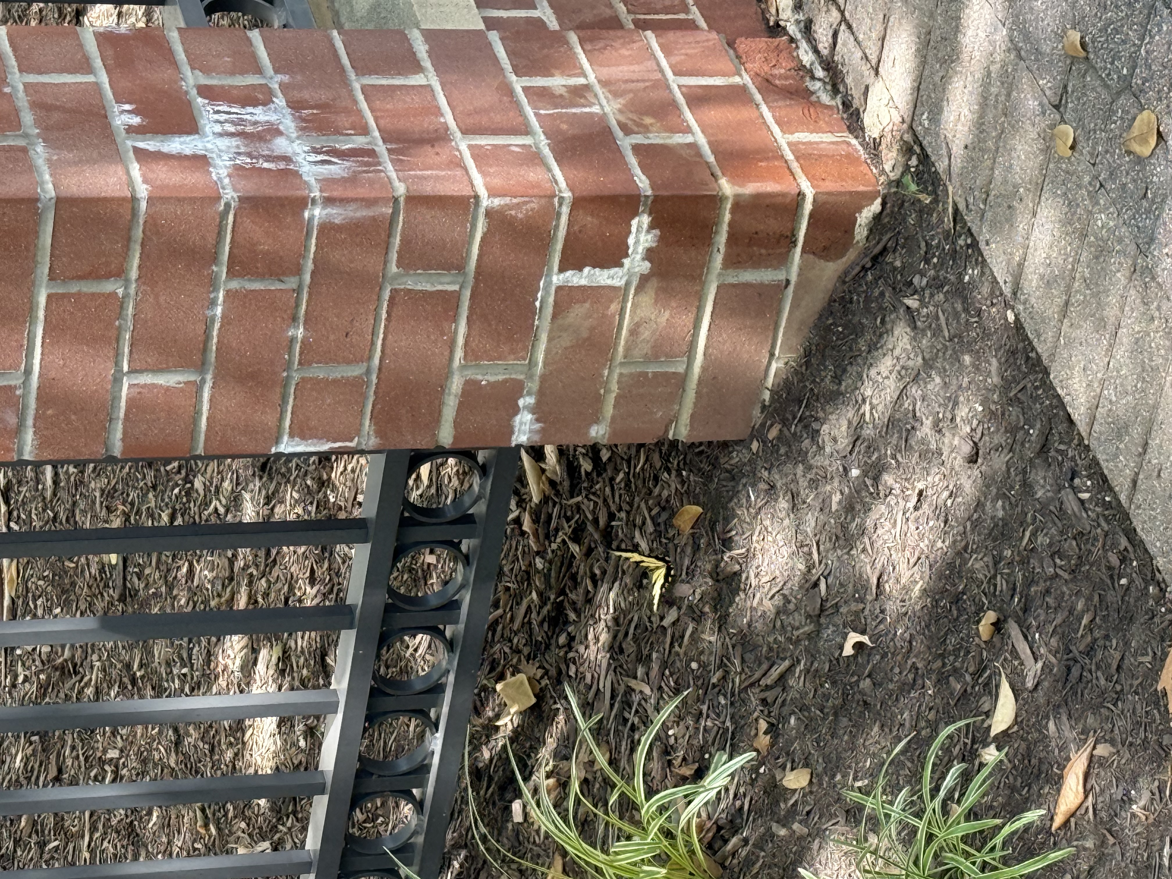 The image features a brick column that supports a wrought iron gate. The column has a few scuff marks, possibly from wear over time, suggesting it has stood the test of time. The iron gate has a sleek design, with circular patterns that add a decorative touch. Surrounding the column is a bed of dark mulch with some scattered leaves and a few green plants, which brings a sense of life to the scene.

Although the image doesn't depict a human subject, one could humorously imagine the column as a grumpy old guardian, weary from standing still all day, while the gate serves as its stylish sidekick. The background’s lush greenery subtly complements the foreground, creating a serene atmosphere. There's no specific cultural reference or event noted, but the combination of brick and iron evokes a classic architectural charm typical in many neighborhoods.