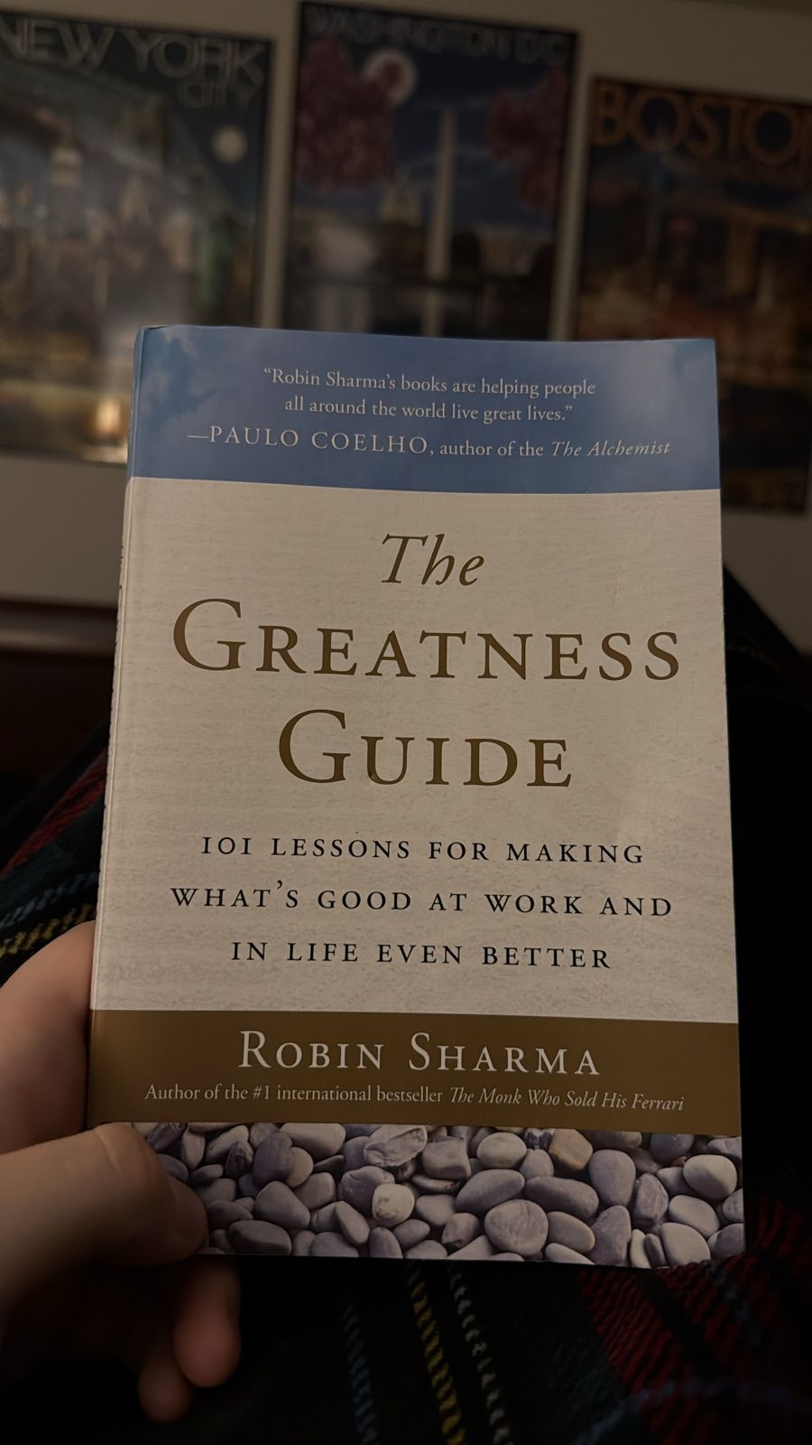 The image features the cover of "The Greatness Guide," a self-help book by Robin Sharma. The book emphasizes 101 lessons aimed at enhancing both work and personal life. The cover is elegantly designed, with a soothing color palette that includes whites and soft tones, complemented by a texture resembling smooth pebbles at the bottom. The subject's demeanor reflects a sense of contemplation and eagerness to improve, as the hand holding the book suggests engagement and interest.
In the background, there are posters of iconic cities, like New York and Boston, hinting at a setting that inspires exploration and growth. This ties into the book's theme of striving for greatness in various aspects of life. The overall context suggests a cozy reading environment, inviting the viewer to reflect on their own journey of self-improvement, sprinkled with a touch of humor as the hand seems to cradle wisdom like a precious gem.