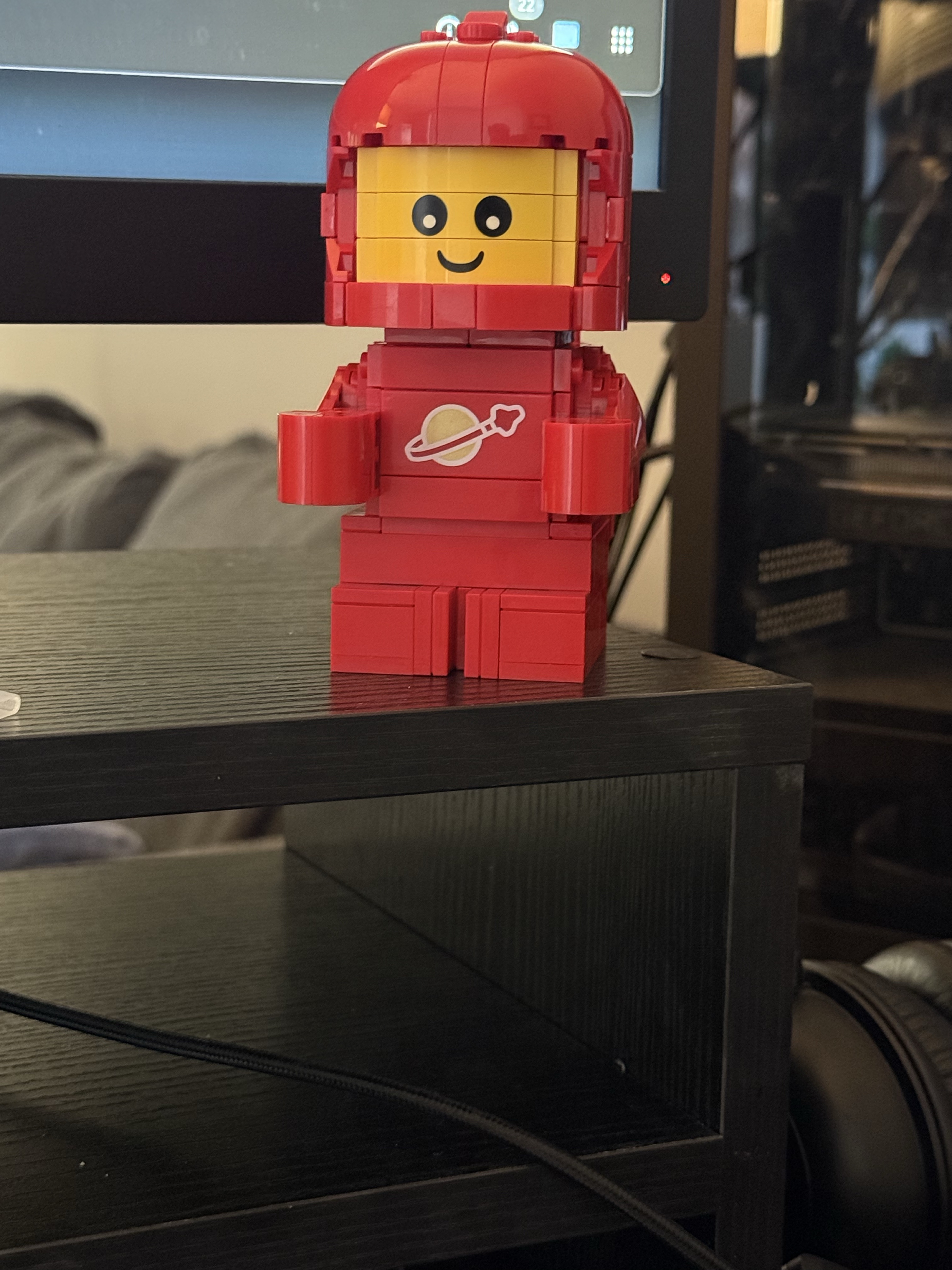 The image features a cheerful LEGO Space Figure, dressed in a bright red spacesuit with a playful smile. Its head is adorned with a matching red helmet, accentuating its enthusiasm for space adventures. The minifigure stands confidently on a black shelf, showcasing its vibrant colors against the dark background. 

In the backdrop, a computer monitor flickers, hinting at a creative workspace filled with potential for imaginative play. The setting suggests a blend of work and leisure, where the boundaries of reality and fantasy blur. 

The LEGO figure, with its iconic planetary logo, embodies the spirit of exploration, perhaps waiting for its next mission to conquer the cosmos—or at least the desk! Its demeanor is inviting, almost as if it's ready to share tales of interstellar escapades with anyone who stops by. The whole scene radiates a lighthearted, whimsical charm, perfect for sparking creativity.