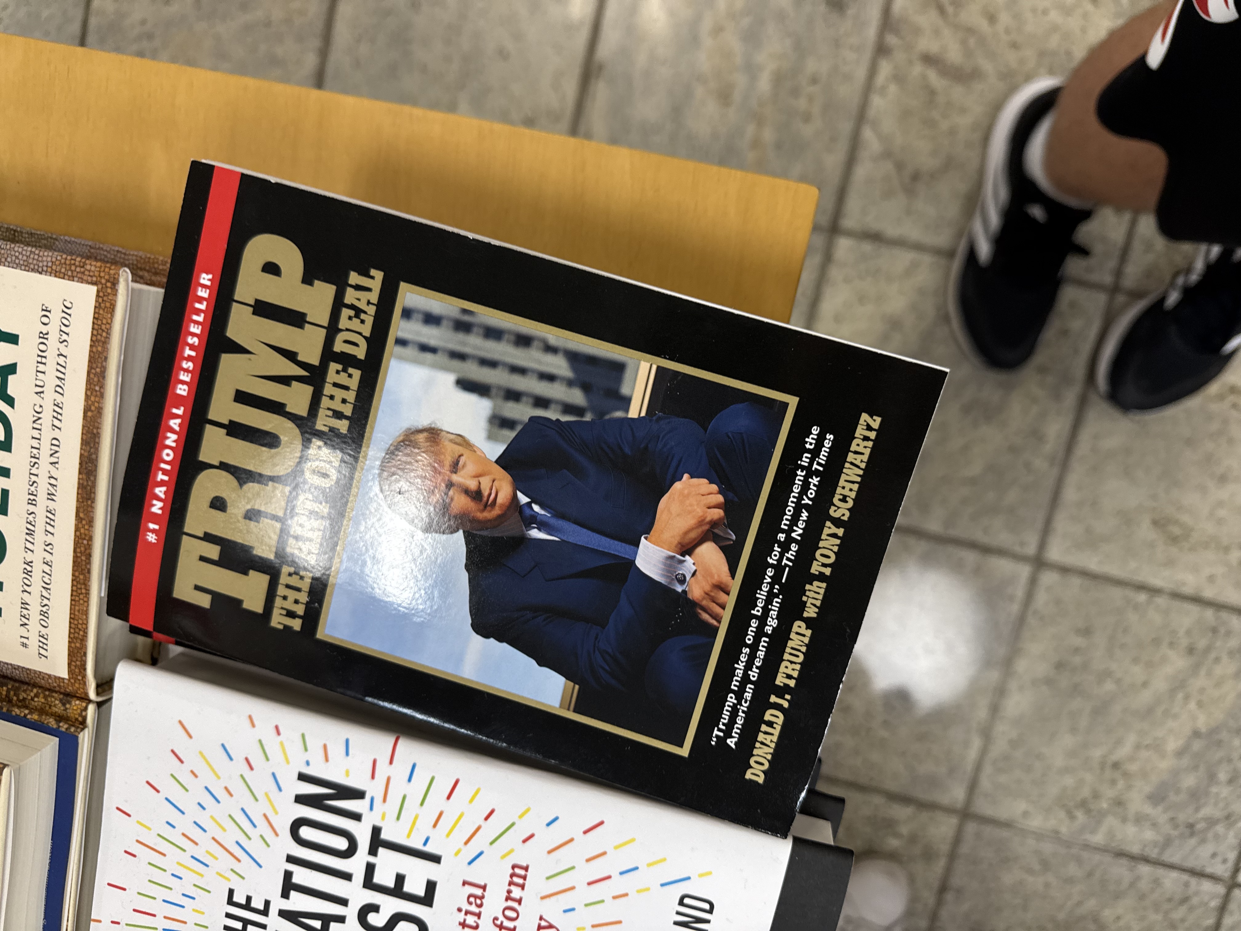 The image features the book cover of "The Art of the Deal" by Donald J. Trump, showcasing a polished, confident Trump in a suit, seated with arms crossed. His demeanor suggests authority and self-assuredness, indicative of his branding as a successful businessman.

In the background, the book is placed among other titles, hinting at a casual bookstore setting. The surrounding books, with colorful covers, create a vibrant contrast to the more muted tones of Trump's book, emphasizing its prominence as a "#1 National Bestseller."

Humorously, one might think that while Trump is sealing deals, the other books are awkwardly trying to compete for attention, perhaps whispering, "Hey, we have great stories too!" Overall, the image captures a blend of business acumen and the casual nature of browsing literature.
