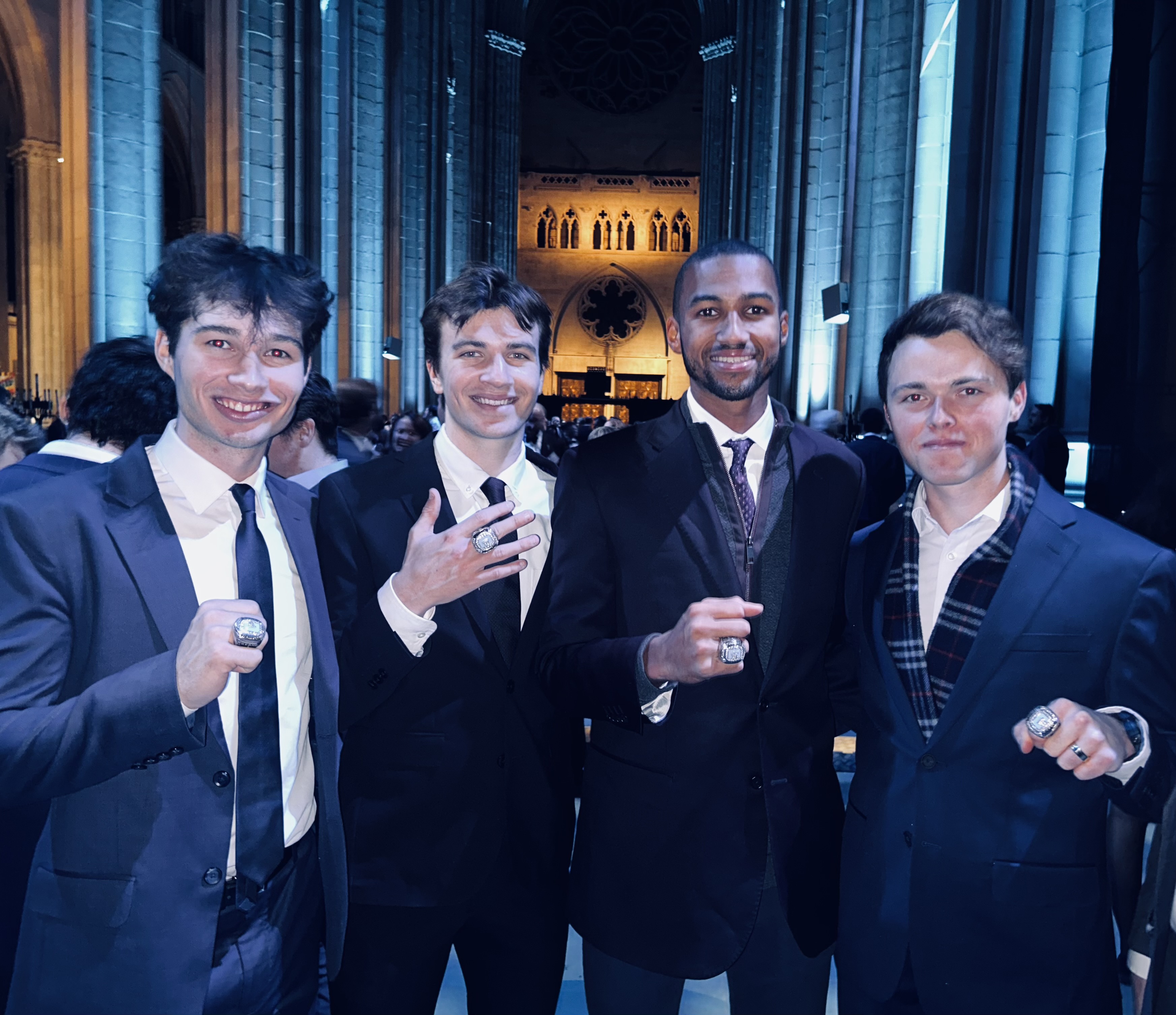 In the image, four young men stand together, each wearing stylish suits and flashing big smiles. Their demeanor exudes confidence and joy, likely celebrating a significant achievement, as indicated by the large rings they are showcasing. The setting appears to be a grand venue, with tall, illuminated stone walls and a soft blue light enhancing the atmosphere, hinting at a formal event or celebration.

The background complements the subjects, suggesting an elegant occasion—perhaps a gala or award ceremony. The architecture suggests a historic or prestigious location, further elevating the significance of their gathering.

Humorously, one might imagine them as a modern-day "Lord of the Rings" fellowship, ready to embark on a quest for the ultimate trophy, but instead, they’re just enjoying the perks of their success!