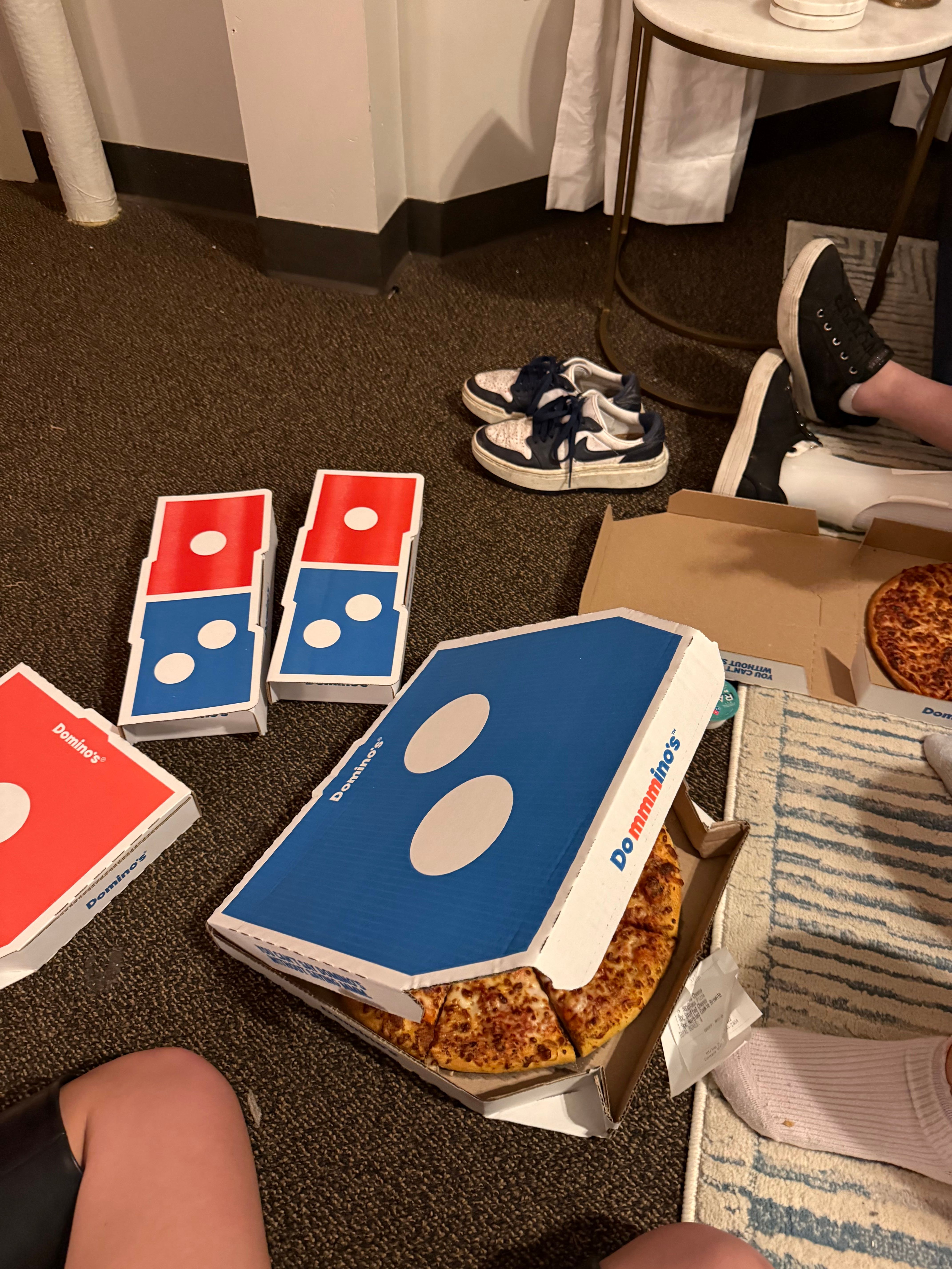 This image captures a quintessential casual gathering, likely a pizza night, with several Domino's Pizza boxes scattered across a dark brown carpet. Two boxes are open, revealing delicious, cheesy pizzas, one of which is humorously tilted, as if mid-grab. A pair of stylish white and navy Nike shoes rests casually nearby, alongside another person's black shoes and white-socked foot, indicating a relaxed, comfortable atmosphere where guests are likely sitting on the floor. The visible legs of two individuals further emphasize this laid-back demeanor. The background, featuring a simple white wall, a curtain, and a small marble-topped table, reinforces the informal indoor setting, perfectly complementing the foreground's scene of shared indulgence and easygoing camaraderie.