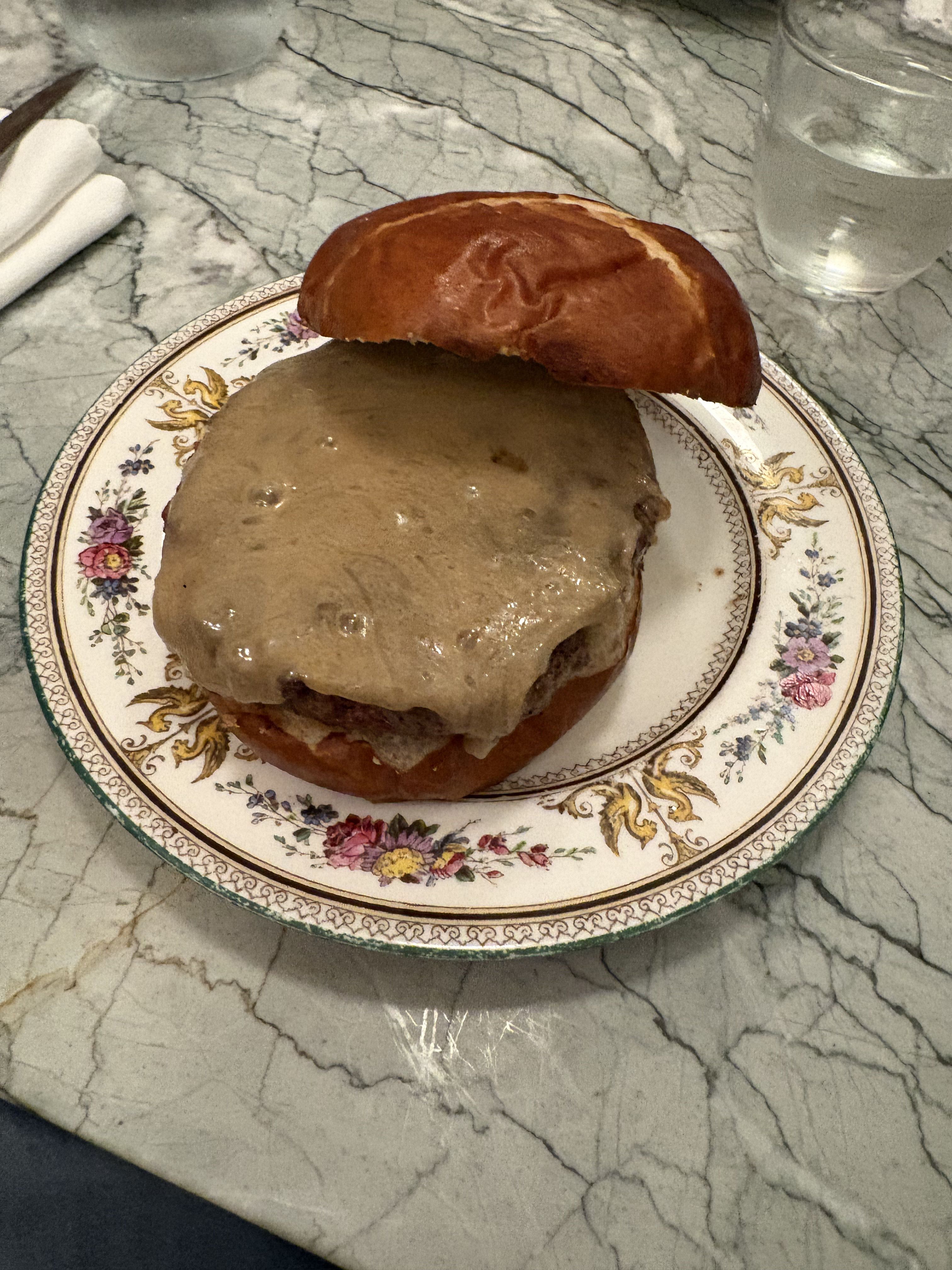 This image features a rather bashful burger, its rich brown pretzel bun slightly tilted as if caught mid-thought. A generous blanket of melted, bubbly cheese drapes over the patty, giving it a wonderfully gooey, almost sleepy demeanor, as if it's just woken up from a delicious nap. It rests on an elegant, floral-patterned plate with golden bird motifs, a humorous contrast to its humble, hearty nature. The background is a sophisticated grey marble tabletop, hinting at a refined dining experience where even a burger gets the royal treatment. Nearby, a glass of water and neatly rolled white napkins await. I am unable to identify any famous or recognizable content in this image, such as celebrities, politicians, popular memes, or specific scenes from known media. The burger seems to be silently daring you to take the first bite.