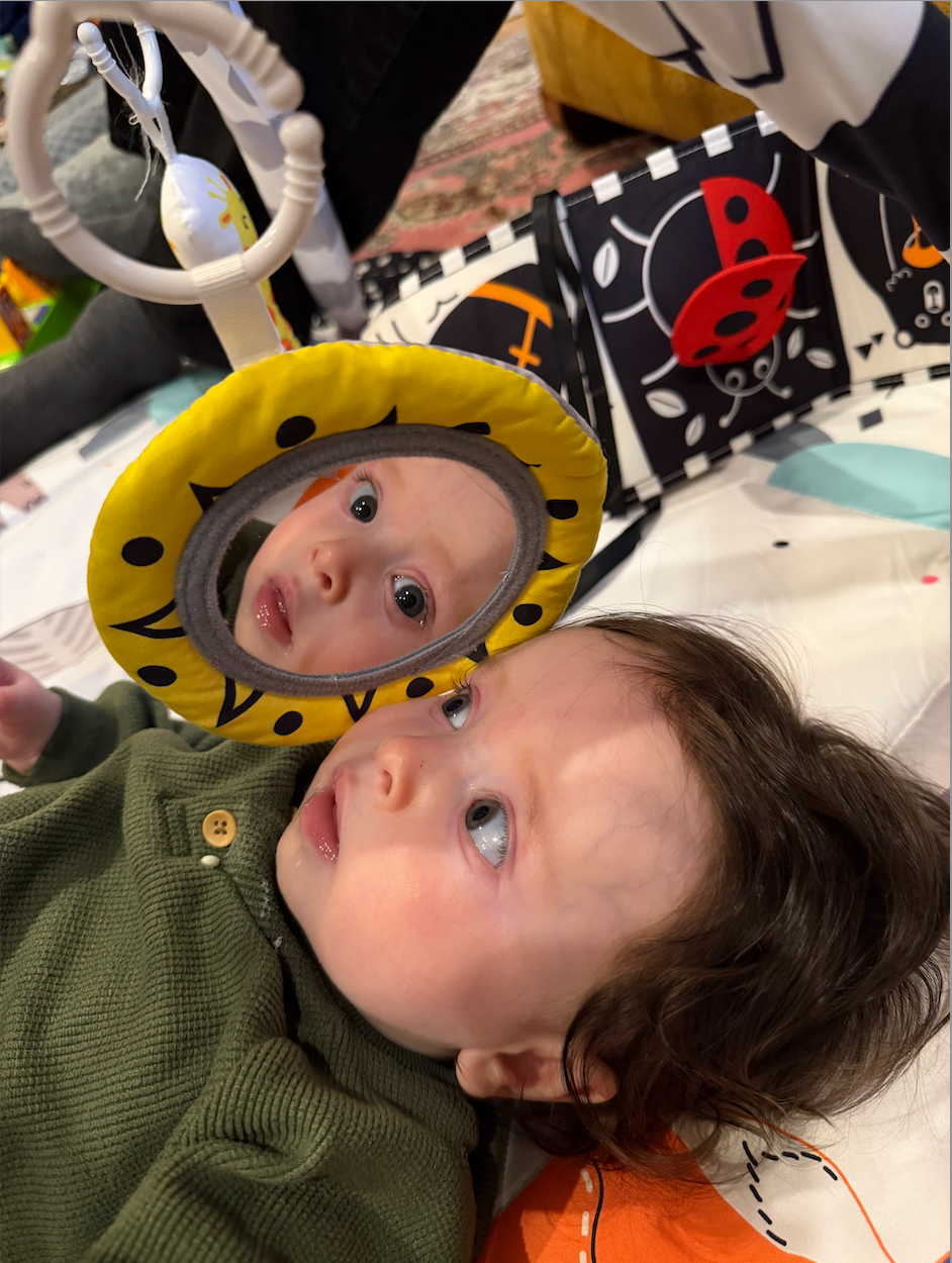 In this charming image, a baby lies on a colorful play mat, gazing intently at their reflection in a round, yellow-framed mirror. The child's demeanor is curious and slightly pensive, with big blue eyes reflecting wonder and innocence. Their soft, curly hair frames their face, and they're dressed in a cozy green sweater, adding to the warmth of the scene.

The background features vibrant toys and a busy play mat, suggesting a playful environment filled with sensory stimulation. The playful designs, including ladybugs and geometric shapes, enhance the joyful atmosphere. This setting captures a moment of self-discovery, where the baby is likely enchanted by their own reflection, leading to humorous thoughts of them pondering, "Who is this adorable stranger?" This delightful image encapsulates the innocence of childhood and the joys of exploration.