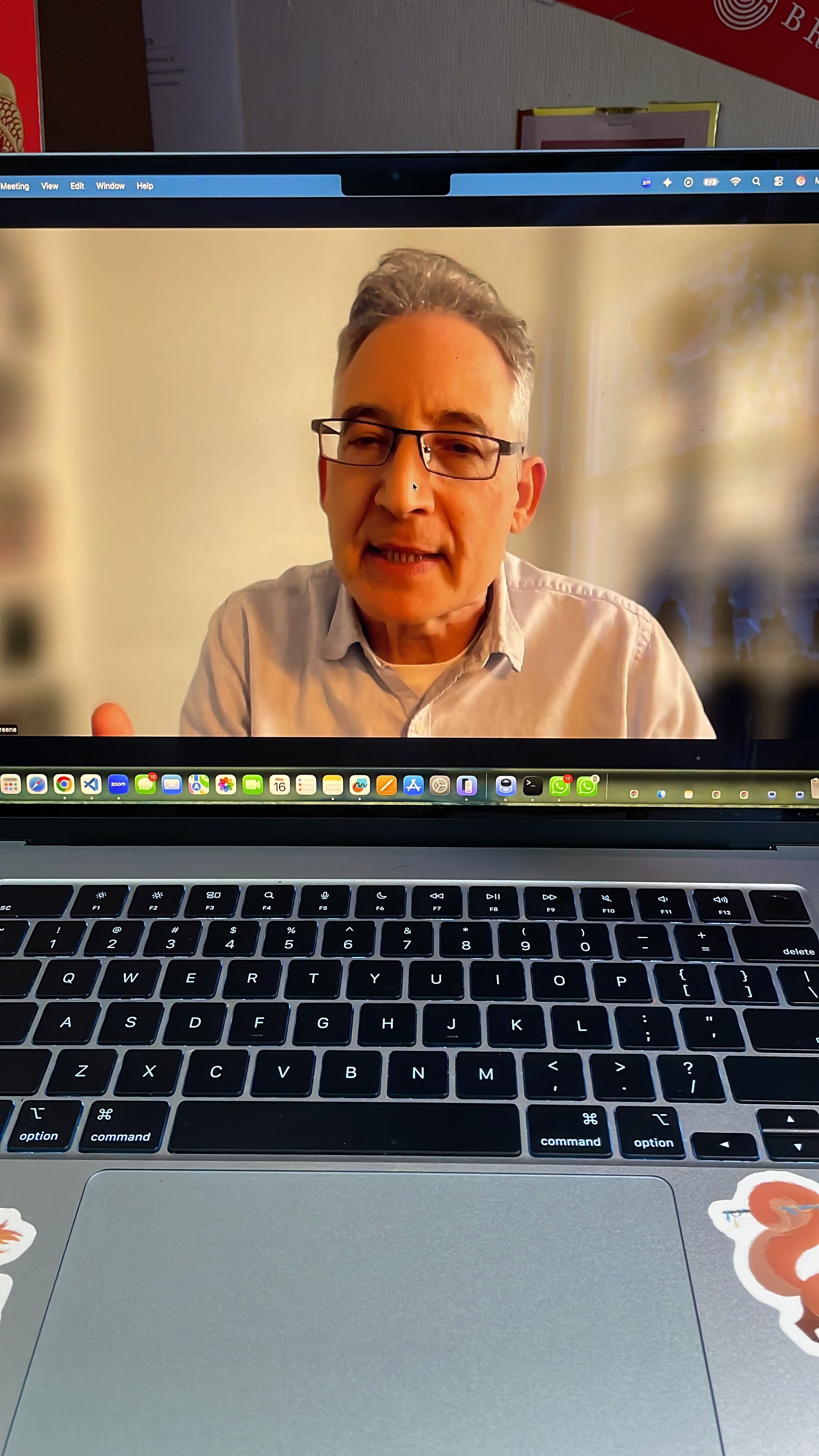 This image shows a generic video call on a laptop, featuring a middle-aged man with glasses and a light shirt. He appears earnest and engaged, mid-sentence with his mouth slightly open and a hand raised, seemingly explaining a point. His focused demeanor is humorously contrasted by a mouse cursor comically resting on his nose. The background on the screen is a plain, light-colored wall, typical of a home or office. The laptop's keyboard and trackpad, with subtle stickers, dominate the foreground, while a blurry real-world background behind the laptop hints at a desk with papers, connecting the digital interaction to its physical setting.
