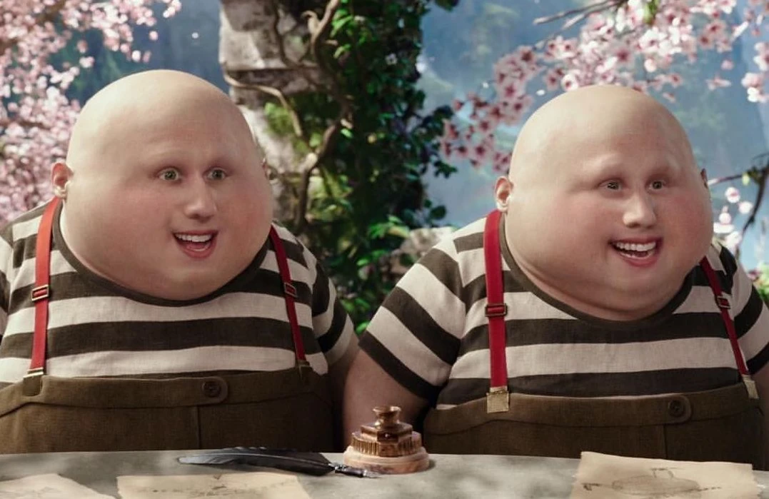 This image features Tweedledee and Tweedledum, famously portrayed by Matt Lucas in the "Alice in Wonderland" live-action movies. Both characters are depicted with their signature bald, round heads and cheerful, wide smiles, exuding a jovial and slightly mischievous demeanor. They wear striped shirts, brown overalls, and bright red suspenders. In the foreground, a quill and an inkwell sit on a table with scattered papers, suggesting they might be engaged in some whimsical paperwork. The background is a vibrant, fantastical landscape characteristic of Wonderland, with lush greenery and delicate pink flowering trees, perfectly setting the scene for their peculiar antics. Their exaggerated features and synchronized, happy expressions add a humorous touch to this iconic duo.