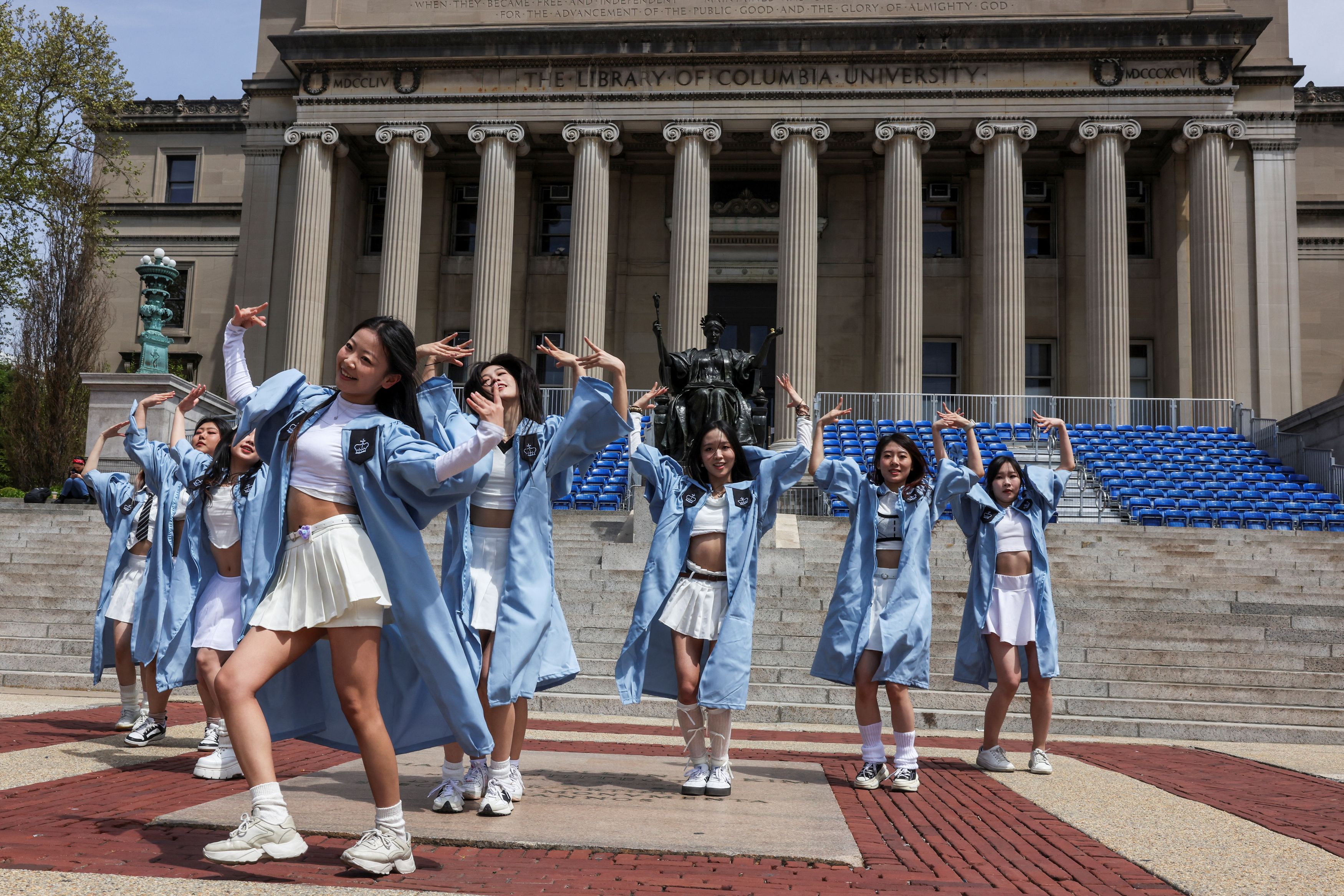 The image showcases a lively dance performance in front of the iconic library of Columbia University. A group of individuals, dressed in light blue robes and white outfits, exudes joy and celebration. Their energetic poses and expressive movements suggest they are celebrating a significant milestone, likely a graduation event.
The backdrop of the university's grand library, known for its impressive architecture and historical significance, adds a prestigious context to the scene. The statue of a seated figure, possibly representing a scholarly figure or a guardian of knowledge, stands as a silent witness to their celebration.
Humorously, it looks like the dancers are trying to outshine the library itself, turning this serious academic setting into a vibrant dance floor! Their playful spirit contrasts sharply with the solemnity typically associated with such a revered institution, making for a delightful visual spectacle.