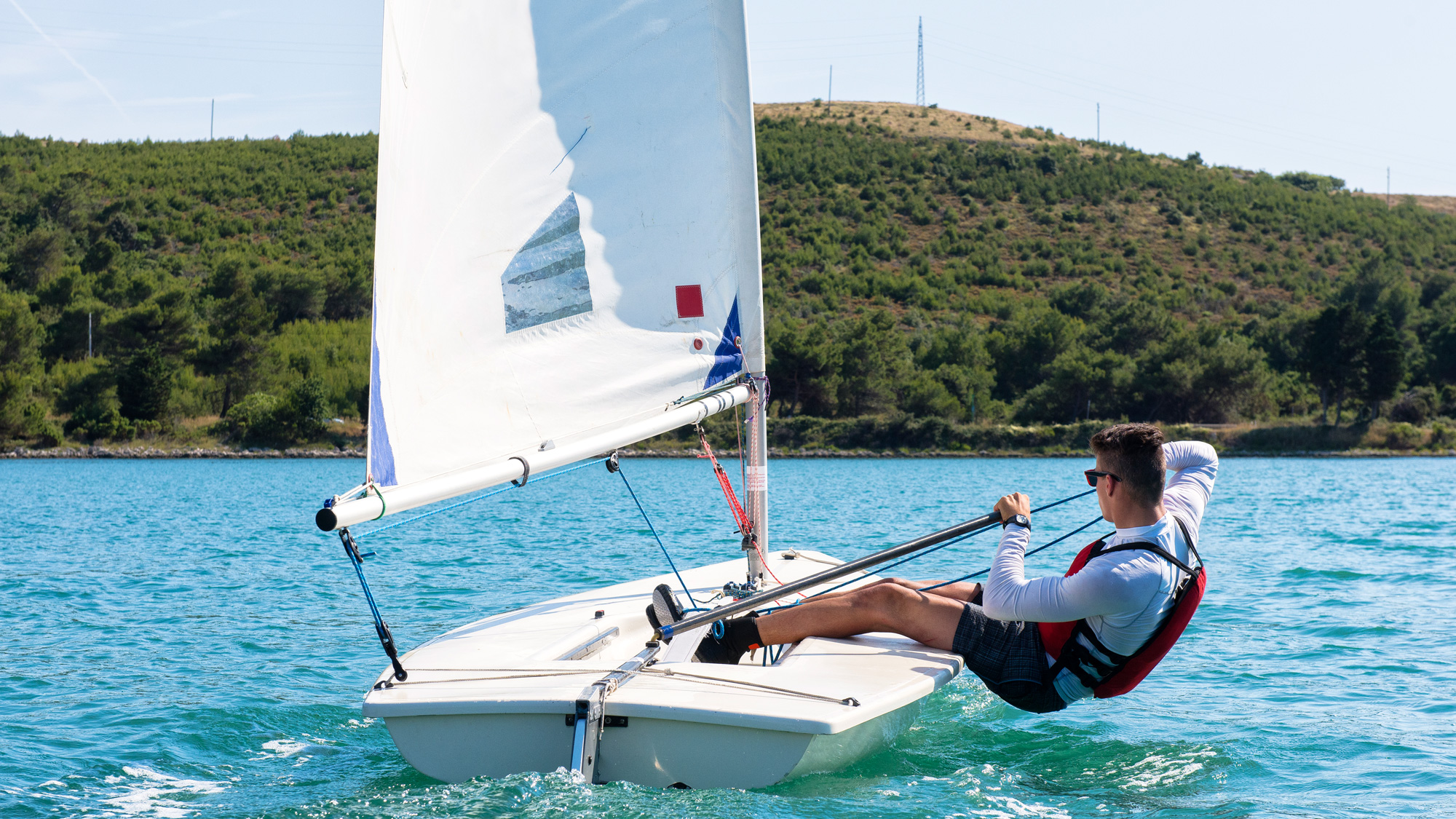 In this vibrant image, a young man is sailing confidently on a small boat, embodying a sense of adventure and exhilaration. His relaxed posture, with one leg propped up and sunglasses on, suggests he's enjoying a sunny day on the water. The light blue water contrasts beautifully with the lush green hills in the background, creating a serene yet dynamic atmosphere.
The gentle ripples around the boat emphasize the movement and freedom of sailing, while the white sail catches the wind, symbolizing exploration. The background, featuring a hillside dotted with trees, adds depth and a sense of tranquility, suggesting a perfect escape from the hustle and bustle of everyday life.
This scene captures the essence of summer fun, reminding us that sometimes, the best adventures come from simply being out on the water, soaking in nature's beauty. It's a slice of life that evokes joy and relaxation, making one wish to join in on the sailing escapade!