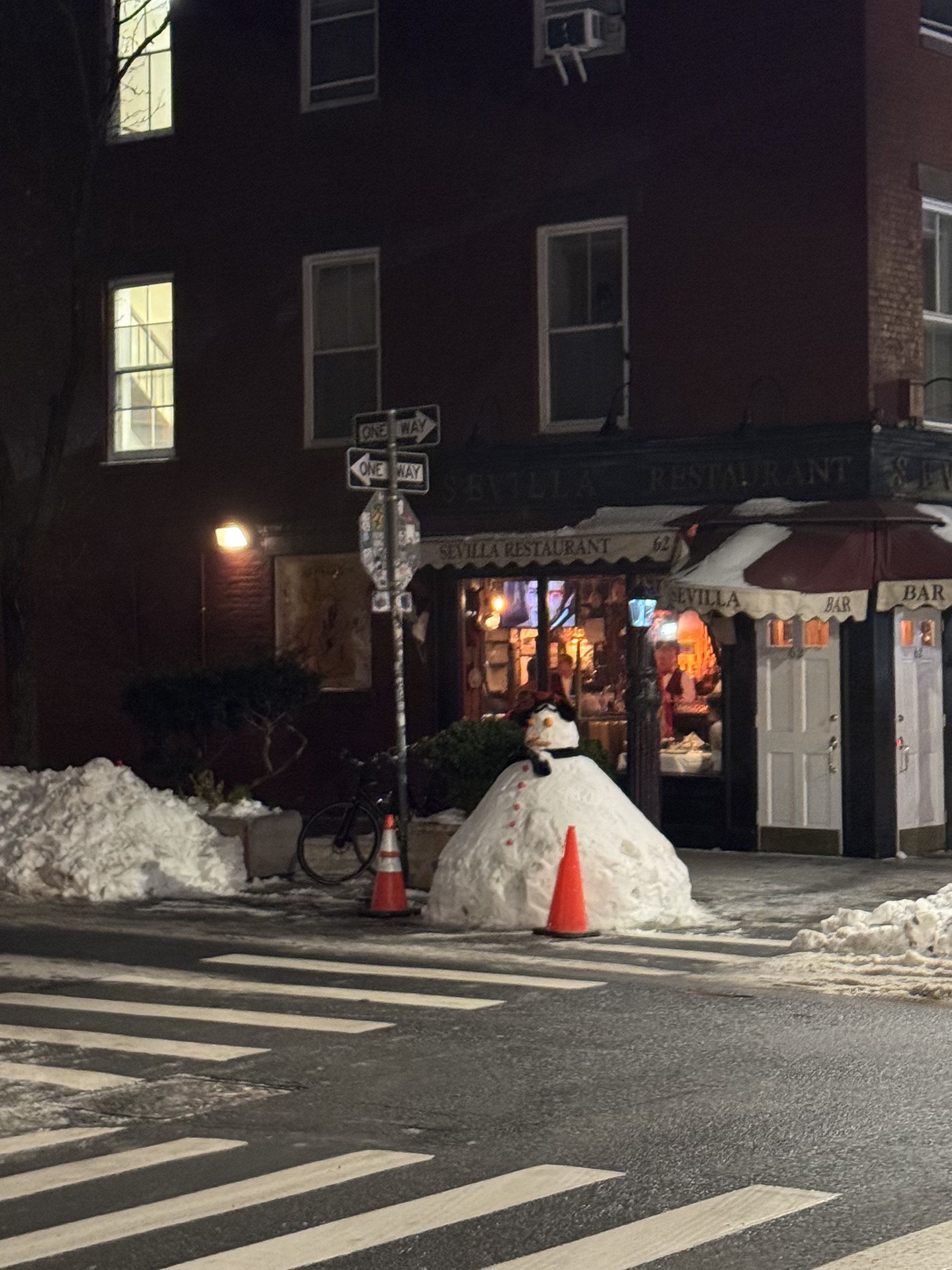 A colossal snowman, looking rather dapper and slightly imposing, stands guard on a snowy urban street at night. Dressed in a black scarf and a dark hat, it appears to be the unofficial bouncer for the "Sevilla Restaurant" and "Bar" behind it, with two orange traffic cones strategically placed as if marking its personal space. The warm, inviting glow from the restaurant's windows contrasts humorously with the chilly, snow-laden foreground, where large piles of snow line the sidewalk. This frosty sentinel, with its button eyes and carrot nose, seems to be patiently awaiting its next patron, or perhaps just enjoying the quiet winter evening in front of this iconic establishment, adding a whimsical touch to the New York City streetscape.