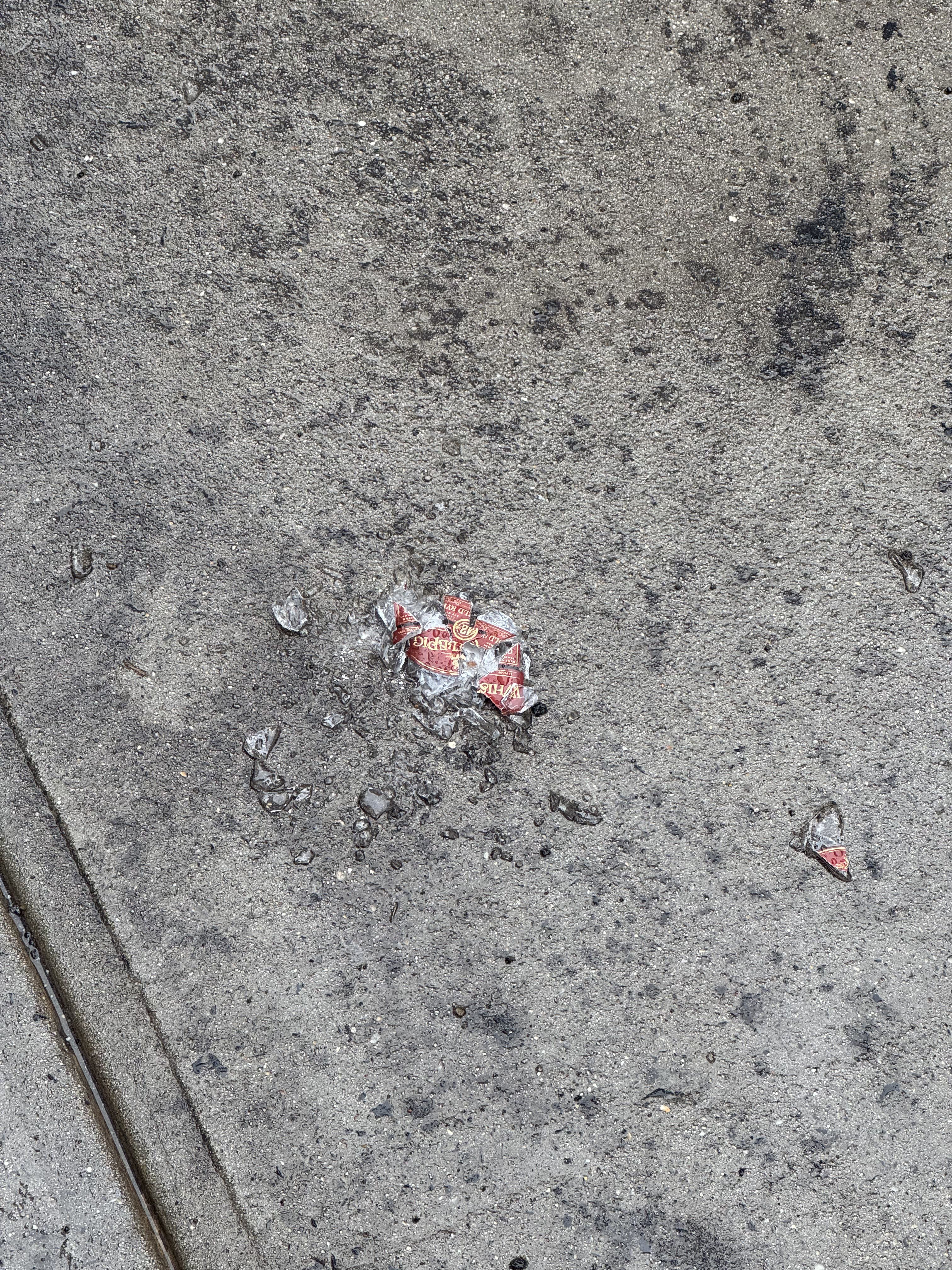 A forlorn, crumpled red and white wrapper, possibly from a candy or snack, lies dejectedly on a rough, wet concrete surface. It appears to be melting into its surroundings, with shards of ice or clear plastic scattered around it like shattered dreams. The wrapper, once a proud vessel of deliciousness, now wears a demeanor of utter defeat, having been unceremoniously discarded and left to brave the elements. Its vibrant red is muted by the dampness, blending into the gritty grey pavement. The background, a mundane sidewalk, perfectly underscores its current, rather unglamorous, existence. It's a tiny tragedy of consumerism, a sweet memory now just a soggy, forgotten mess.
