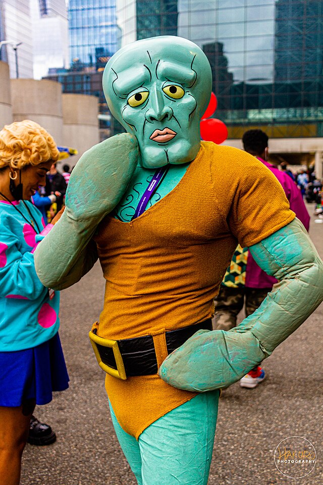 A person in a full-body Handsome Squidward costume from SpongeBob SquarePants strikes a pose, embodying the character's exaggerated, melancholic 'beauty' with a hand on their hip and a flexed arm. The mask perfectly captures his iconic, pouty expression and large yellow eyes, creating a humorous juxtaposition with the muscular physique. To the left, a person partially visible in a costume resembling Boo from Monsters, Inc. smiles, adding to the playful atmosphere. The urban background, featuring modern glass skyscrapers, suggests a public event like a comic convention or cosplay gathering, where these beloved animated characters humorously step out of their fictional worlds into reality. The scene is a vibrant celebration of pop culture.