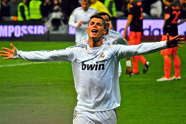 Cristiano Ronaldo, in his iconic white Real Madrid jersey with Adidas and bwin logos, is captured mid-celebration on a vibrant green football pitch. His demeanor is pure ecstasy and triumph, head tilted back, mouth open in a powerful shout, and arms spread wide as if embracing the moment or the entire stadium. The blurred background features other players, some in orange and black, and stadium lights, confirming the intense atmosphere of a professional football match. The partially visible "IMPOSSIR" on an advertising board humorously hints at the 'impossible' feats he often achieves. This image perfectly encapsulates the raw emotion of a goal celebration, a signature moment for the legendary Portuguese footballer.