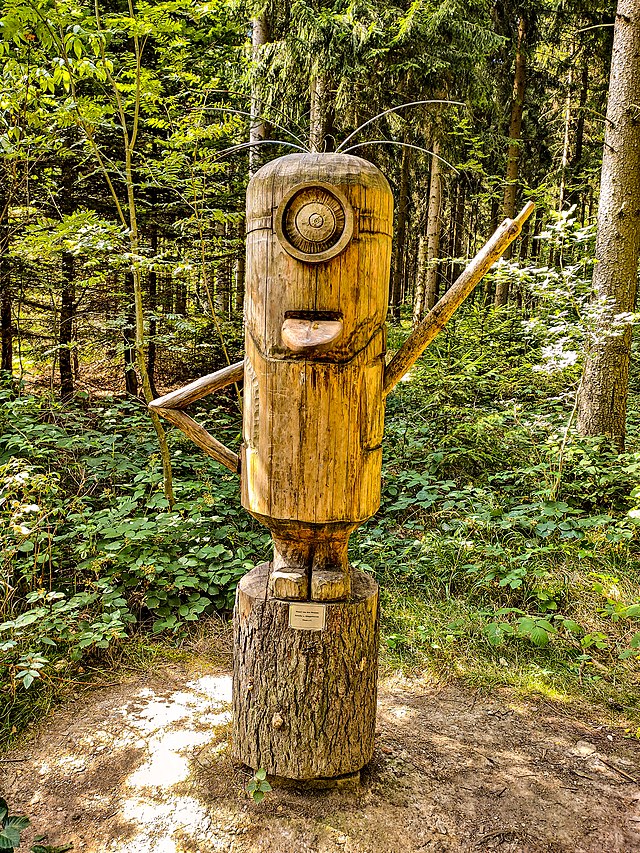 The image features a charming wooden sculpture of a Minion, a beloved character from the "Despicable Me" franchise. The sculpture stands tall in a lush forest, surrounded by vibrant green foliage, which enhances its playful demeanor. With one arm raised in a friendly wave and an exaggerated smile, the Minion seems to invite passersby to join in its whimsical antics.

The natural setting adds to the humor, as this mischievous figure appears to be having a grand time amidst the trees, perhaps plotting its next silly adventure. The contrast between the animated character's playful essence and the serene forest backdrop creates a delightful juxtaposition. It’s as if the Minion has taken a break from its chaotic life to enjoy nature, embodying childlike joy and mischief in a wooden form. The whole scene invites smiles and laughter, making it a perfect spot for a fun photo op.