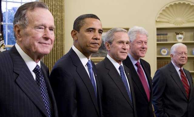 In this iconic image, five former U.S. Presidents—George H. W. Bush, Barack Obama, George W. Bush, Bill Clinton, and Jimmy Carter—stand together in the White House. Their expressions vary from solemnity to slight smiles, reflecting a mix of camaraderie and the weight of their shared responsibility.
The backdrop, with its elegant curtains and bookshelves, emphasizes the historical significance of the gathering, symbolizing continuity in American leadership. Each President, dressed in formal attire, showcases both their unique personalities and the gravity of their roles, while also hinting at the lighter moments that must have been shared off-camera.
The scene captures a rare moment in time, evoking humor in the contrast of their differing styles and eras. It’s almost like a presidential family photo, reminding us that beneath the titles, they are all human, perhaps sharing jokes about past campaigns or political mishaps.