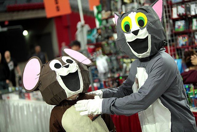 The image features two individuals dressed as Tom and Jerry, the beloved characters from the classic animated series. Tom, the cat, is clad in a gray outfit with exaggerated features, including a wide grin and large, expressive eyes, exuding a playful demeanor. Jerry, the mouse, is in a brown costume, equally cheerful, sporting a big smile that captures the essence of their comedic rivalry.

The background hints at a lively event, likely a convention or festival, with colorful merchandise displays and excited attendees. This setting enhances the playful atmosphere, suggesting a celebration of nostalgia and pop culture. The humorous dynamic between the characters, combined with their cartoonish costumes, invites laughter and joy, reminiscent of their iconic chases. The scene encapsulates the fun spirit of cosplay, bringing these charming characters to life in a whimsical way.