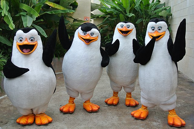 The image features four cheerful mascot costumes of the iconic Penguins of Madagascar: Skipper, Kowalski, Rico, and Private. They stand in a line, all with wide, open-mouthed smiles and expressive, wide eyes, conveying an enthusiastic and welcoming demeanor. Each penguin has at least one flipper raised in a wave, as if greeting an audience. The background consists of lush, vibrant green tropical foliage and a light-colored building, strongly suggesting a theme park, zoo, or resort setting, which perfectly complements the animated characters' adventurous, exotic origins. The scene humorously captures the penguins' signature playful and slightly mischievous personalities, ready for their next mission or photo opportunity.