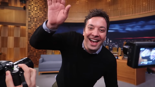 In this lively image from "The Tonight Show Starring Jimmy Fallon," the host, Jimmy Fallon, beams with a wide smile while waving enthusiastically. His demeanor radiates warmth and friendliness, inviting viewers into the moment. He’s dressed casually in a black sweater over a light blue shirt, embodying a relaxed yet polished vibe typical of late-night television.

The background features a cozy set with warm wood paneling and a cityscape backdrop, suggesting the familiar atmosphere of a talk show. A couple of cameras are pointed at him, emphasizing the dynamic interaction with the audience and the lively nature of live television.

This scene captures the essence of entertainment and spontaneity, with Fallon’s playful energy reminiscent of a humorous moment where he might be about to share a funny story or engage in a light-hearted banter. It's a snapshot of joy, making it a delightful moment for fans of the show.