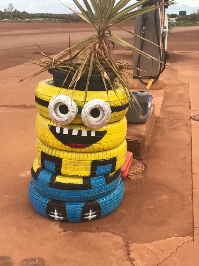 This cheerful, DIY Minion, crafted from a stack of painted tires, stands proudly as a roadside attraction. Its wide, toothy smile and large, foil-covered eyes exude a goofy, welcoming demeanor, perfectly capturing the essence of the beloved characters from the Despicable Me franchise. A spiky plant sprouts from its head, comically serving as its wild, unkempt hair. This resourceful art installation, painted in iconic yellow and blue overalls, brightens the dusty, red-earthed setting of what appears to be a gas station. The juxtaposition of the whimsical Minion planter against the utilitarian gas pump and arid landscape adds a touch of unexpected humor and charm, making it a delightful, if slightly out-of-place, guardian of the pumps.