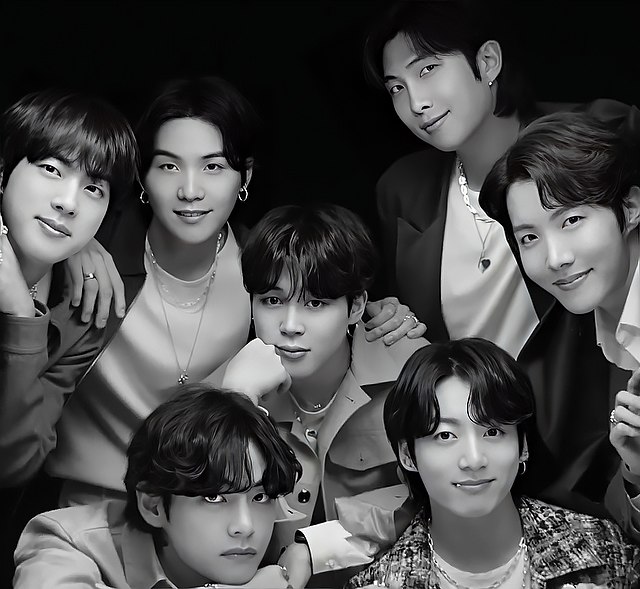 This striking black and white portrait features all seven members of the globally renowned K-pop group BTS, exuding their signature charisma. From left to right, top row: Jin, Suga, RM, and J-Hope offer warm, inviting smiles. Below them, Jimin rests his chin thoughtfully, while V delivers an intense, captivating gaze, contrasting playfully with the more relaxed expressions. Jungkook, at the bottom right, also shares a gentle smile. The stark black background serves to dramatically highlight each member, emphasizing their individual yet cohesive presence. It's a classic studio shot, likely for promotional material, where their collective star power is the undeniable focus. The humorous element lies in the sheer concentration of visual appeal, almost overwhelming the viewer with their collective charm and varied, yet equally captivating, demeanors.