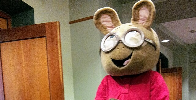 The image features a cheerful Arthur Read mascot costume, the beloved aardvark from the "Arthur" children's animated television series. Arthur is depicted with a wide, open-mouthed smile, his iconic round glasses perched on his nose, and wearing his signature red long-sleeved shirt. His demeanor is undeniably friendly and inviting, perfectly capturing the character's wholesome nature. The background consists of light-colored walls and a prominent wooden door or paneling, suggesting an indoor setting like a school, library, or event hall. This mundane background humorously contrasts with the larger-than-life cartoon character, implying a public appearance or meet-and-greet event for fans of the show. The slightly grainy quality of the image adds to its candid, event-photo feel.