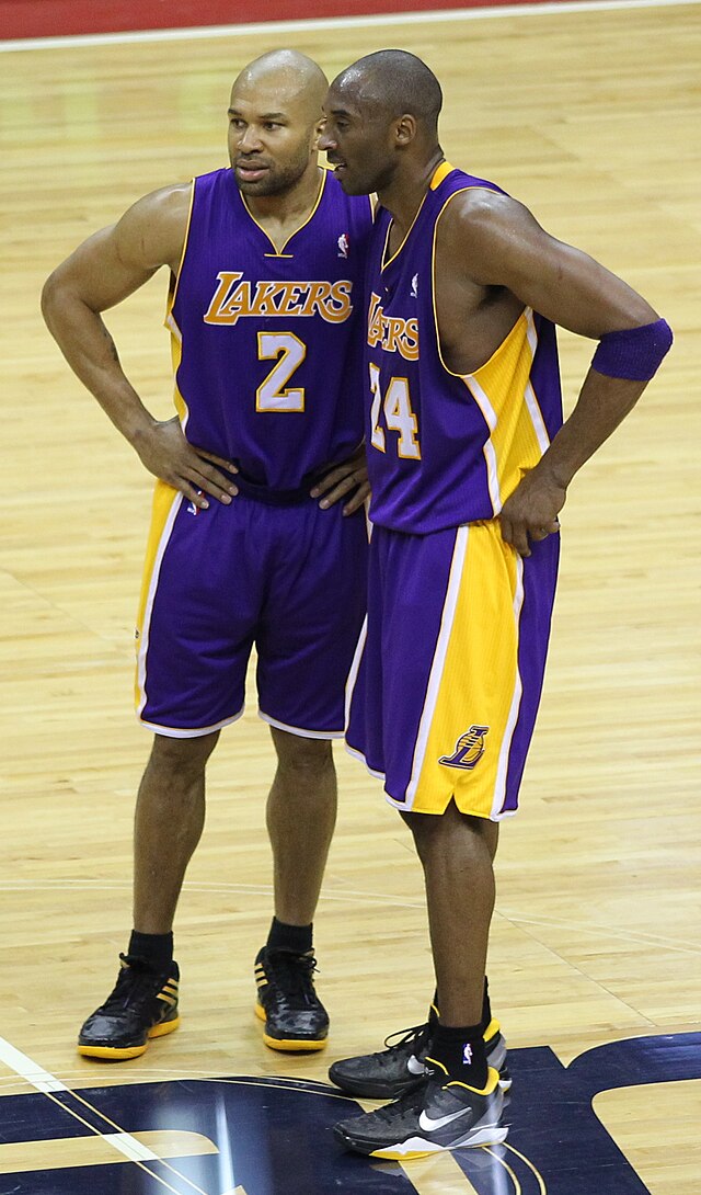 The image captures two iconic NBA players, Kobe Bryant and Derek Fisher, in a moment of teamwork during a game, both sporting their distinctive purple and gold Los Angeles Lakers uniforms. Kobe, wearing number 24, leans slightly closer to Derek, who wears number 2, suggesting a serious discussion or strategic planning. Their body language conveys focus and camaraderie, indicative of their strong partnership on the court.
The background features a polished wooden basketball court, typical for NBA games, emphasizing the competitive environment. While there's no specific landmark visible, the court itself is a symbol of the Lakers' storied history.
In this scene, the duo's demeanor reflects a blend of intensity and mutual respect, showcasing the essence of teamwork. It's as if they are plotting an elaborate play, ready to dazzle fans with their skills. The overall atmosphere is charged with anticipation, embodying the spirit of basketball.