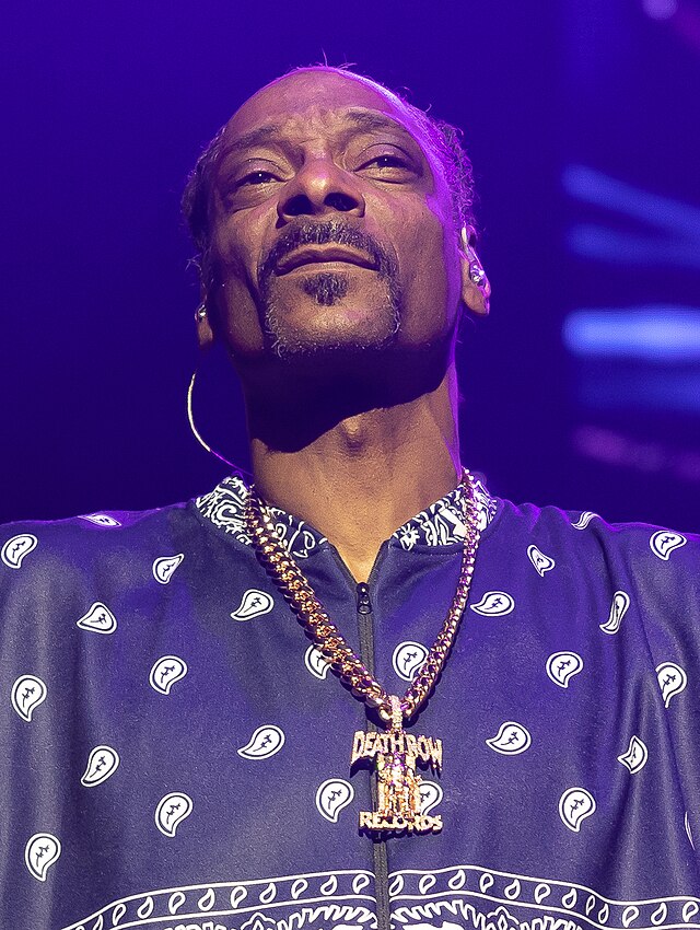 Snoop Dogg is captured mid-performance, looking upwards with a contemplative yet confident demeanor, bathed in dramatic purple stage lighting. He sports a dark blue jacket adorned with a white paisley bandana pattern, a clear nod to his iconic style. A prominent, gleaming gold chain with a diamond-encrusted "Death Row Records" pendant hangs around his neck, powerfully referencing his legendary history with the hip-hop label. The dark, illuminated background emphasizes his presence on stage, creating an intimate concert atmosphere. It's almost as if he's checking if the smoke machine is working overtime, or perhaps just admiring the venue's ceiling decor.