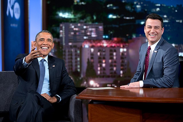 The image captures a jovial moment between former President Barack Obama and host Jimmy Kimmel on the set of "Jimmy Kimmel Live!". Both men are laughing heartily, with Obama gesturing animatedly, suggesting a particularly humorous exchange. Obama, dressed in a dark suit and blue tie, sits comfortably, while Kimmel, in a grey suit and red tie, leans forward slightly at his desk. The background features a stylized cityscape at night, a common backdrop for late-night talk shows, emphasizing the urban, entertainment-focused setting. The genuine mirth shared by a former world leader and a comedian creates a wonderfully lighthearted and relatable scene, highlighting the relaxed atmosphere of the show.