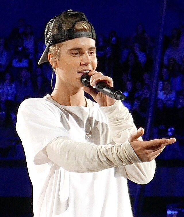 In this vibrant concert scene, Justin Bieber captivates the audience with a dynamic performance. His demeanor is engaging and animated, as he holds a microphone with one hand while extending the other towards his fans, fostering a sense of connection. Dressed in a casual white long-sleeve shirt and a black cap, he embodies the youthful and laid-back vibe that characterizes his style.
The background features an enthusiastic crowd, their faces illuminated by stage lights, highlighting the energy of the live event. This setting emphasizes the connection between the performer and his audience, integral to the concert experience.
Humorously, one might imagine Bieber's outstretched hand as an invitation for fans to join him in a spontaneous dance-off—who wouldn’t want to showcase their moves alongside a pop icon? The scene perfectly encapsulates the joy and excitement of live music, making it a memorable moment for all involved.