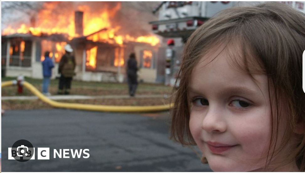 The iconic "Disaster Girl" meme captures a young girl with a mischievous, almost smug smirk, looking directly at the viewer. Her demeanor is calm and knowing, creating a humorous juxtaposition with the dramatic background: a house fully engulfed in roaring flames. Firefighters and onlookers are visible, observing the blaze, with a fire truck partially seen on the right. The background directly provides the ironic context, implying the girl might be responsible for the fiery spectacle or is simply enjoying the chaos. Her subtly sinister smile amidst the destruction is the core humorous element, making this image a widely recognized cultural phenomenon.