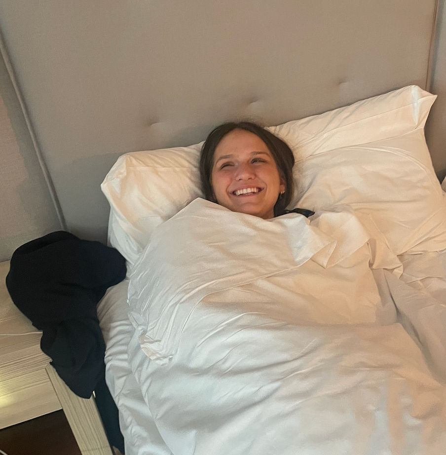 A young woman is captured mid-laugh, beaming with pure joy while nestled deeply in a pristine white bed. Her eyes sparkle as she looks upwards, perhaps at a playful companion or a funny screen. She's completely enveloped in the soft white duvet, resembling a contented human burrito, with only her head peeking out. Her dark hair frames her radiant face, and small gold hoop earrings add a subtle gleam. The light grey upholstered headboard provides a soft backdrop, while a dark hoodie casually draped over a bedside table with a warm lamp suggests a cozy, personal bedroom setting. The scene exudes comfort and uninhibited happiness, a perfect snapshot of a relaxed, humorous moment. I do not recognize any famous or recognizable content in this image.