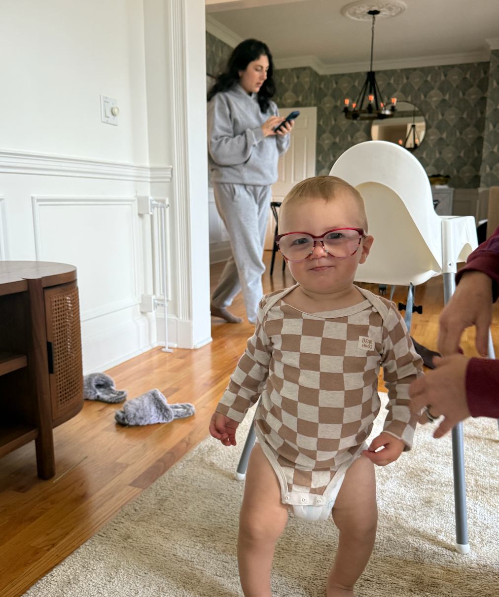 A hilariously serious toddler, sporting oversized red-framed glasses, stands front and center with a comically determined expression, as if contemplating profound thoughts. Dressed in a brown and beige checkered onesie, this little intellectual seems to be taking their first wobbly steps, possibly aided by hands just out of frame. In the background, a casual home scene unfolds: a woman in grey loungewear walks by, engrossed in her phone, seemingly oblivious to the tiny genius in the making. Fluffy grey slippers lie abandoned on the polished wooden floor, hinting at a relaxed domestic setting. The patterned wallpaper and chandelier in the distance suggest a stylish yet lived-in home. The baby's earnest gaze through the ill-fitting spectacles is the highlight, creating a wonderfully humorous and candid moment of everyday life.