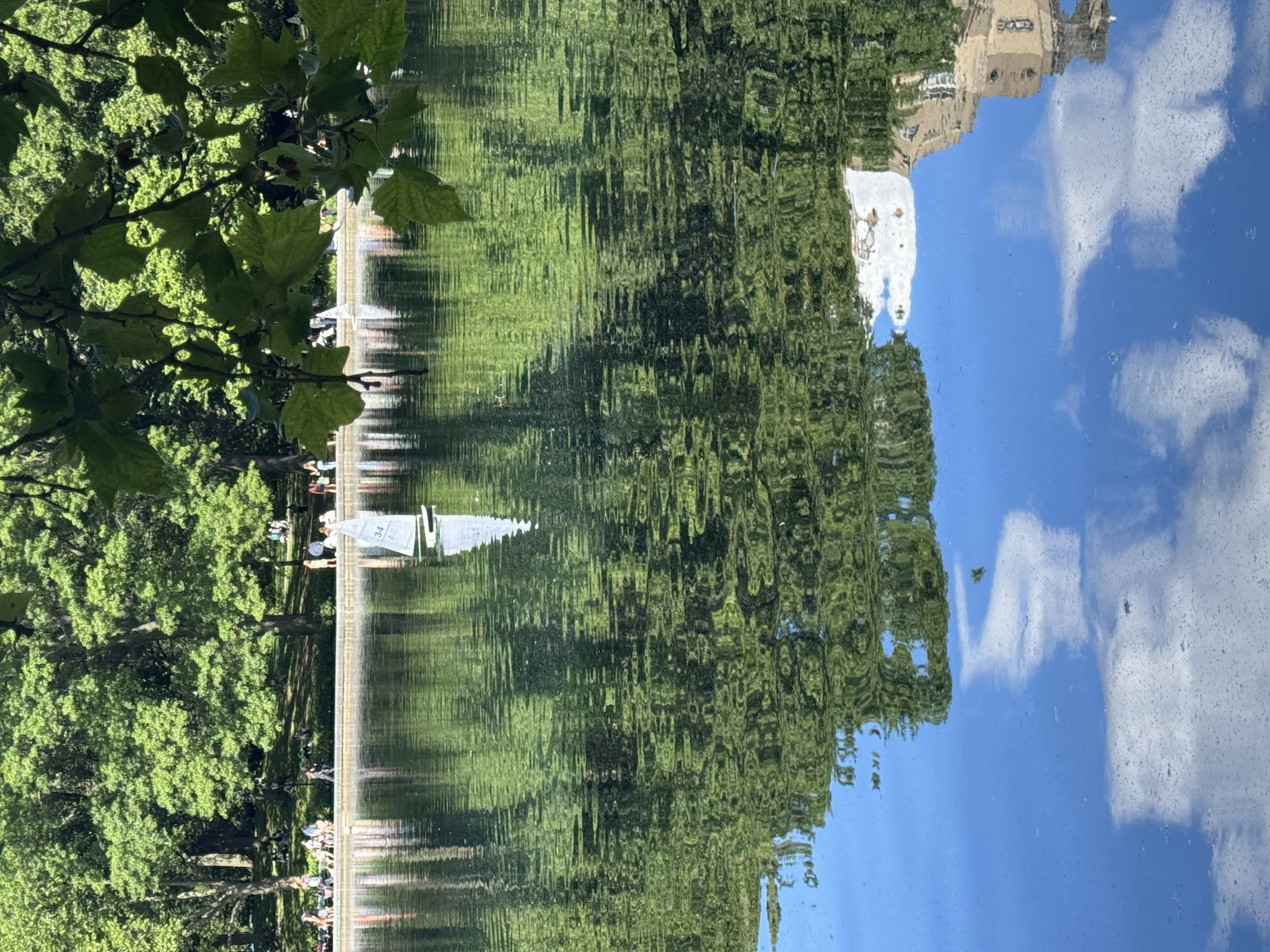 The image captures a serene outdoor scene, featuring a tranquil body of water reflecting lush greenery and a bright blue sky dotted with clouds. A small sailboat, white with its sails raised, glides gracefully across the water, creating a picturesque contrast against the vibrant greens and blues. The reflections in the water are beautifully distorted, adding an artistic flair to the scene.

In the background, people can be seen enjoying the day, perhaps picnicking or strolling along the water’s edge, contributing to a relaxed and joyful atmosphere. The surrounding trees frame the image, their leaves casting dappled shadows on the water, enhancing the peaceful setting.

This could evoke a sense of nostalgia reminiscent of idyllic summer days, making one feel as though they’re part of a delightful outdoor adventure. The overall mood is light-hearted, inviting laughter and enjoyment in nature.