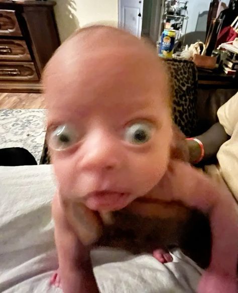 The image features a baby with exaggeratedly large eyes and a quirky expression, creating a humorous and endearing demeanor. The child appears to be in motion, with a slightly blurred focus that adds to the playful chaos of the moment. 

In the background, there are hints of a cozy living space, including wooden furniture and a patterned rug, suggesting a warm, familial setting. The presence of everyday items like a can on a shelf hints at the casual nature of the scene.

This kind of image often evokes laughter and joy, reminiscent of viral memes where cute babies steal the spotlight with their expressive faces. The overall vibe is lighthearted, capturing a fleeting moment of childhood wonder that many can relate to.