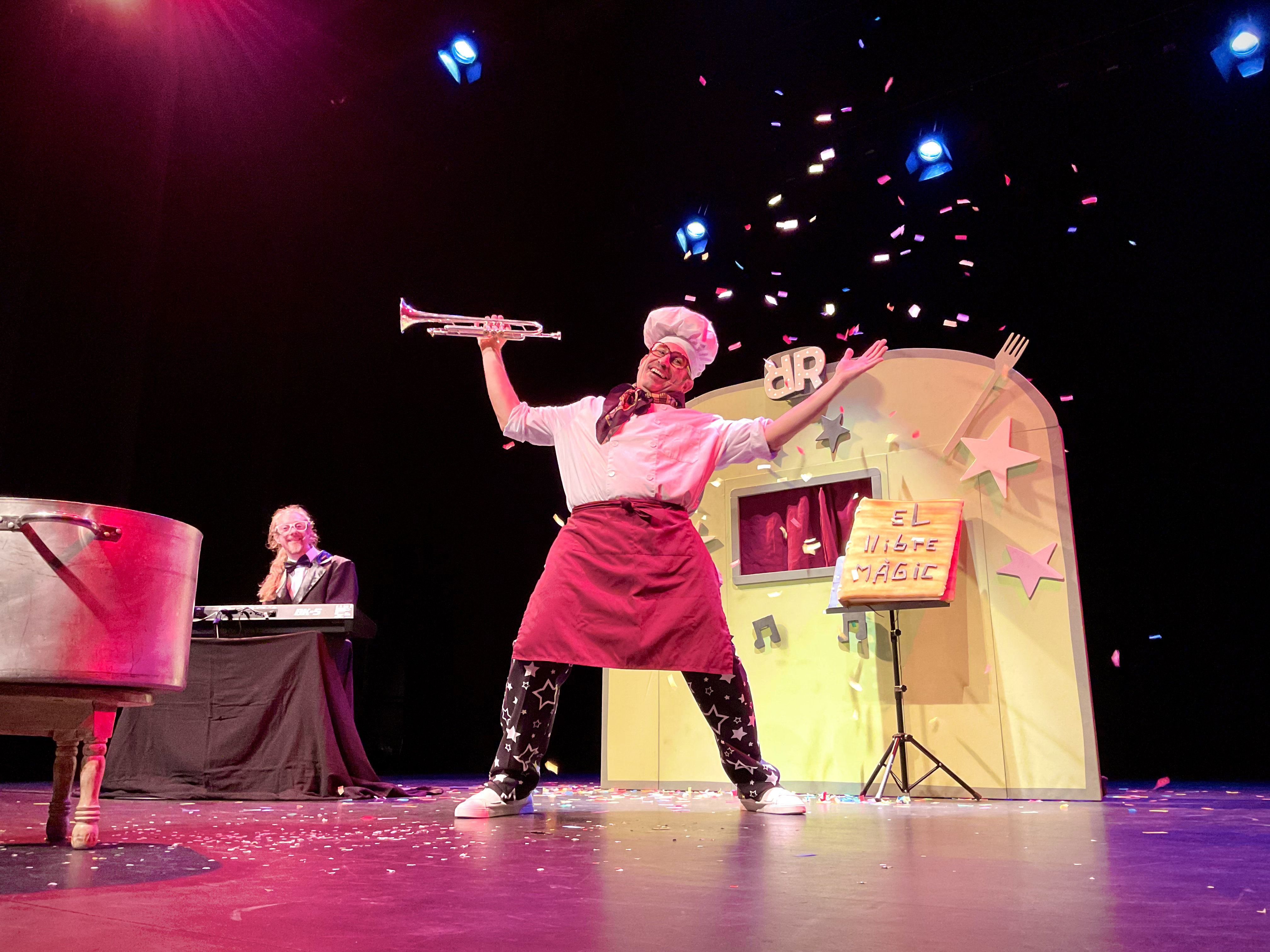 A vibrant stage performance features a joyful man dressed as a chef, complete with a tall white hat, red apron, and star-patterned pants. He holds a trumpet triumphantly overhead, beaming with an energetic and theatrical demeanor as colorful confetti rains down around him. In the background, a whimsical yellow prop resembling a stylized kitchen or food truck, adorned with pink stars and a sign reading "EL LLIBRE MAGIC" (The Magic Book), sets a playful scene. To the left, another person with long blonde hair and glasses, dressed in a tuxedo, smiles while playing a keyboard, adding to the festive atmosphere. The entire setup suggests a lively, humorous, and perhaps magical children's show or theatrical production. I do not recognize any famous or recognizable content in this image.