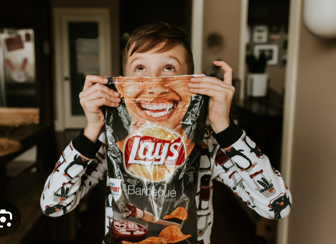 This delightful image captures a young child, sporting charming penguin pajamas, playfully holding a Lay's Barbecue potato chip bag directly in front of their mouth. The bag's printed smiling face perfectly aligns with the child's, creating a hilarious illusion of an enormous, chip-craving grin. The child's eyes, wide with a mischievous glint, gaze upwards, suggesting a moment of playful anticipation or being caught in a joyful act. The warm, softly blurred background, hinting at a cozy home environment with a subtle bunny decoration, reinforces the casual, fun context. This candid shot beautifully conveys a lighthearted demeanor and the simple joy of childhood antics, making the viewer smile at the clever visual gag.