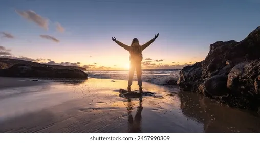 The image captures a person silhouetted against a breathtaking sunset on a serene beach, arms outstretched in a triumphant, joyful pose. Their demeanor suggests immense freedom and elation, perhaps a classic 'King of the World' moment reminiscent of *Titanic*. The vibrant sky, painted with hues of orange, yellow, and blue, perfectly frames the figure, reflecting beautifully on the wet sand. Rocky outcrops on either side anchor the scene, while gentle waves lap at the shore, adding to the tranquil yet powerful atmosphere. The background is integral, enhancing the feeling of liberation and connection with nature. One might humorously imagine them shouting, 'I'm finally on vacation!'
