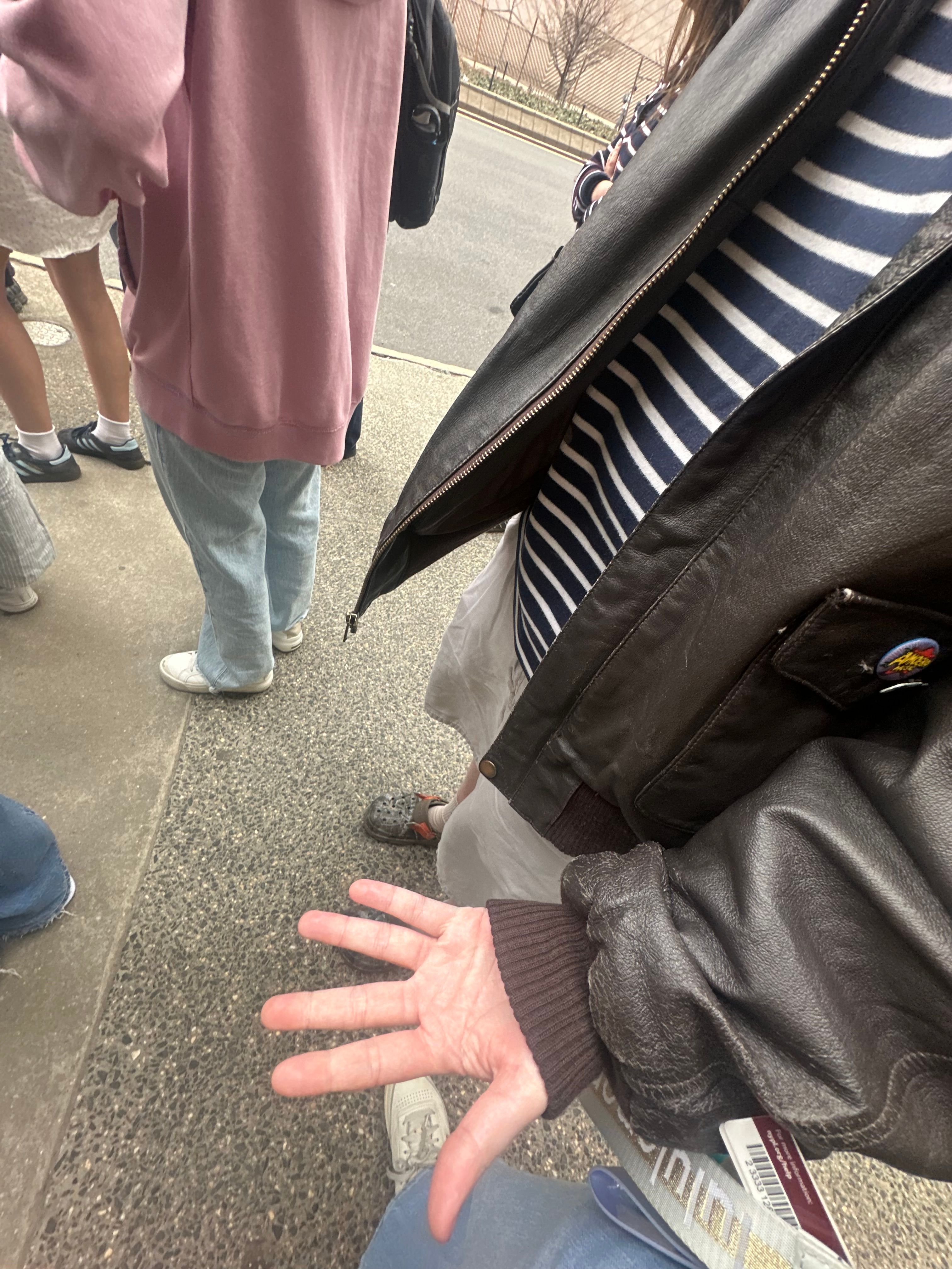 The image captures a casual scene where a group of people is gathered, likely waiting in line. The foreground features a hand extended outward, suggesting a gesture of greeting or perhaps an offer for a high-five. The hand appears relaxed but slightly wrinkled, hinting at age or experience, and is adorned with a cozy, brown cuff from a leather jacket that adds a touch of rugged style to the outfit.
In the background, various individuals are present, dressed in casual attire—a mix of hoodies and light clothing, indicative of a laid-back atmosphere. The setting appears urban, with a sidewalk leading to a blurred street, hinting at a lively environment.
Overall, the image conveys a sense of camaraderie among friends, perfectly encapsulating a moment of anticipation or lightheartedness typical of a day out. The scene feels relatable and evokes a humorous image of the hand possibly trying to get attention in a crowd.