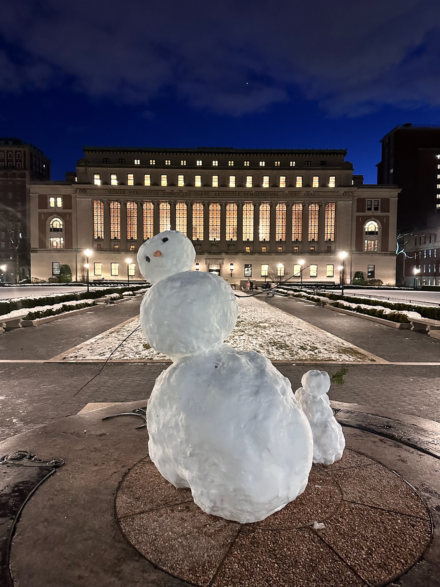 The image features a whimsical snowman prominently placed in front of the grand Columbia University building at night. The snowman, with its round body and cheerful expression—complete with a carrot nose and twig arms—exudes a playful demeanor. Behind it, the university's historic architecture is beautifully illuminated, showcasing tall windows that reflect a warm glow against the cool night sky.

The serene winter scene contrasts the academic prestige of Columbia University, suggesting a delightful fusion of scholarly pursuits and seasonal joy. The snowman almost seems to stand guard at this iconic institution, inviting students and passersby to join in the winter festivities.

Humorously, one might imagine the snowman contemplating the meaning of life, perhaps pondering if it should enroll in a philosophy class given its proximity to the university. After all, what better place to find enlightenment than beside a snowman on campus?