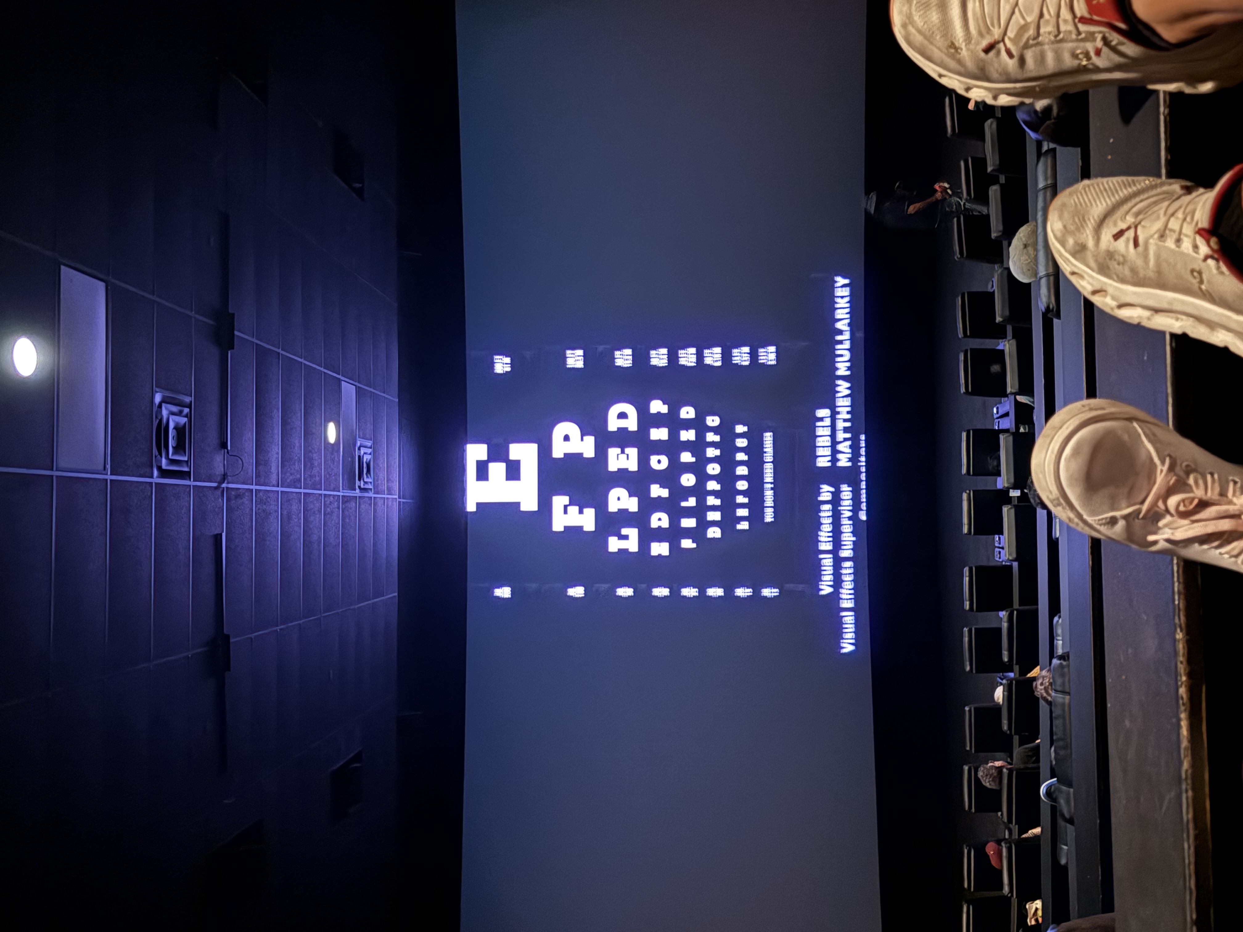 The image depicts a cinema setting where the focus is on a large screen displaying an eye chart, typically used for vision tests. The foreground features a pair of feet, presumably belonging to a viewer relaxing in their seat. The shoes are casual and white, suggesting a comfortable, laid-back demeanor. The overall ambiance is dark, typical of a movie theater, with rows of empty seats visible behind.

The background, showcasing the eye chart, adds a humorous twist, as it humorously contrasts the typical movie scenes one might expect in a theater. Instead of cinematic action, viewers are greeted with a vision test, perhaps hinting at a lighthearted or quirky movie experience ahead.

The text at the bottom credits visual effects personnel, adding a layer of irony as audiences are reminded of the behind-the-scenes work while they await the show. It’s a playful nod to the cinematic experience!