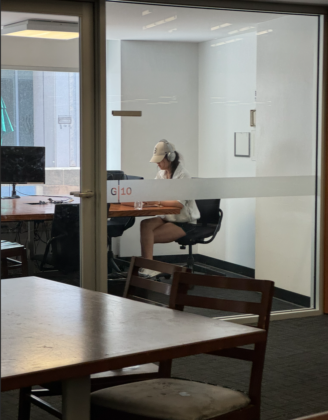 The image captures a focused individual seated at a table in a study room, likely in a library or educational setting. She wears headphones and a cap, appearing deeply engaged in her work, possibly studying or on a video call. Her demeanor suggests concentration, as she leans slightly forward, with a water bottle nearby, a sign of preparation for a long session.

The background features a glass wall, providing a glimpse of additional tables and chairs, hinting at a collaborative study environment. The presence of a computer monitor suggests a tech-friendly space, conducive to modern learning. 

The wooden furniture contrasts with the sleek, minimalistic design of the room, creating a cozy yet professional atmosphere. With the sunlight filtering in, the scene feels inviting, perfect for those seeking both solitude and focus. Overall, it embodies the spirit of student life, where every corner is a potential hub of productivity.