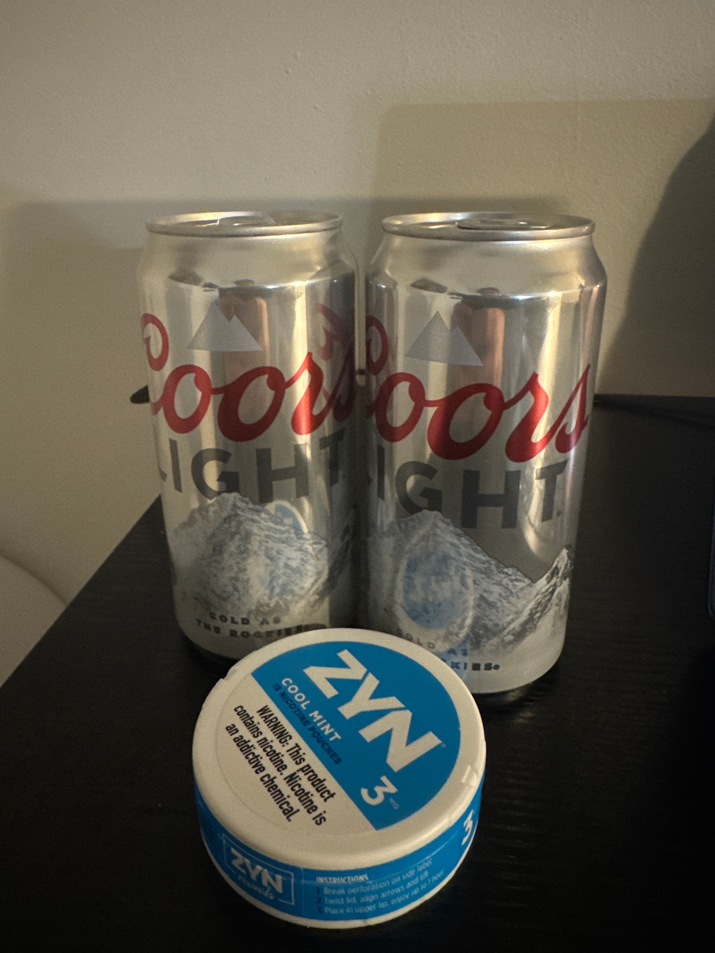The image features two cans of Coors Light, a popular American beer brand, positioned prominently beside a can of ZYN nicotine pouches. The Coors Light cans are silver with bold red lettering, showcasing the brand's iconic mountain logo, suggesting a refreshing and laid-back vibe often associated with social gatherings. The ZYN can, in contrast, boasts a vibrant blue design labeled "Cool Mint," hinting at a modern, tobacco-free alternative that appeals to a younger demographic. 

The overall scene conveys a casual atmosphere, possibly set in a living room or a social space, where these products may be enjoyed together. The background is simple and neutral, allowing the focus to remain on the products. This combination humorously reflects the recreational habits of many, showcasing a friendly rivalry between beer and nicotine alternatives at a gathering, ready to fuel some light-hearted fun!