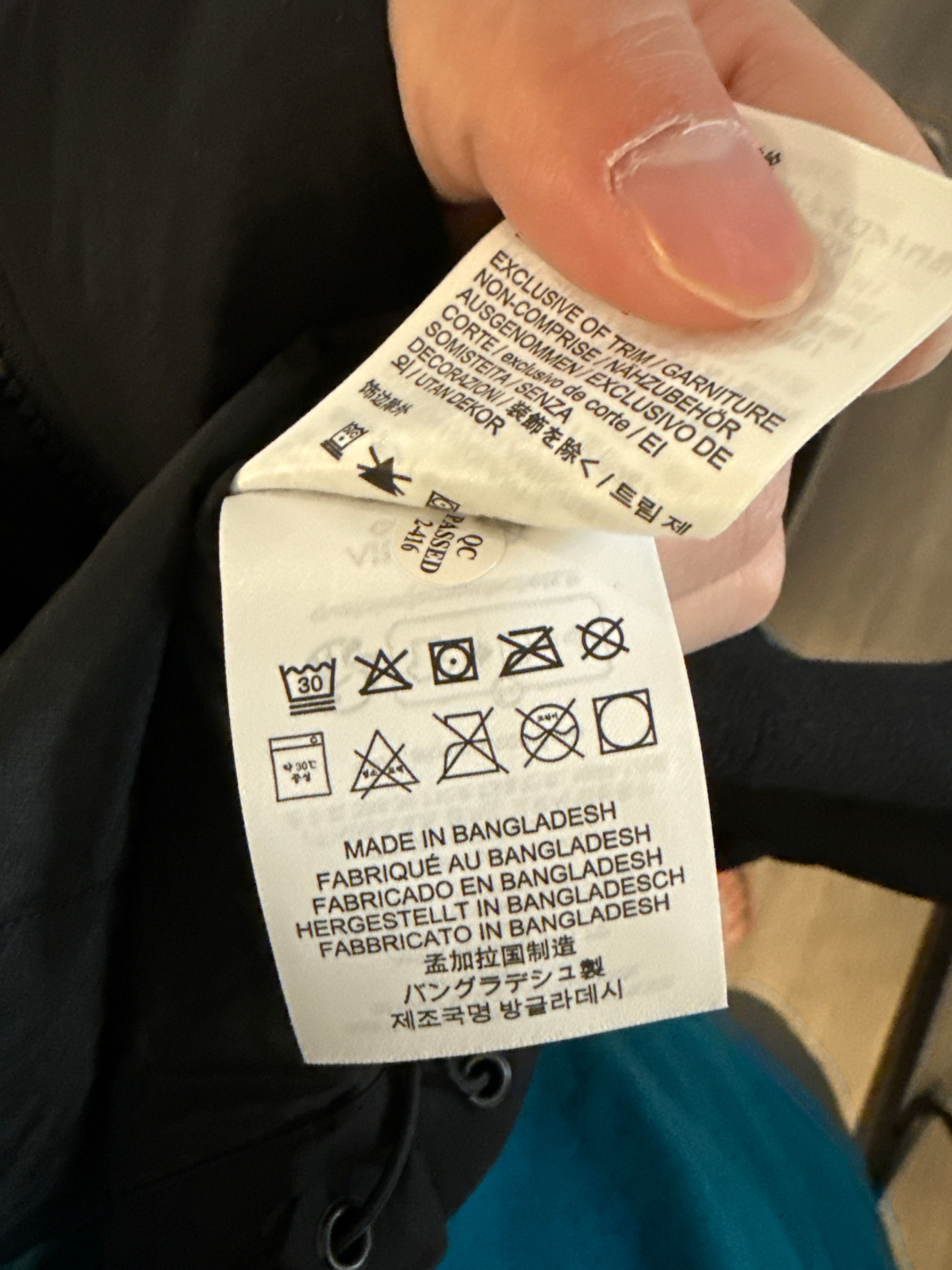 The image features a close-up of a clothing label held in a person's hand. The label is filled with various care instructions, including washing symbols and information about the fabric's origin, specifically noting it was made in Bangladesh. The hand appears to have a light skin tone and shows signs of wear, possibly indicating frequent use or manual work. 

The background is somewhat blurred, but hints at an indoor setting, likely a casual environment such as a home or a store. This suggests a relatable, everyday context where one might be checking clothing care instructions. 

Humorously, one might imagine the label as a wise old sage, imparting crucial life advice on how to treat your garments: 'Do not bleach!' and '30°C wash only!'—the ultimate guardians of fabric care, ready to save you from wardrobe disasters!