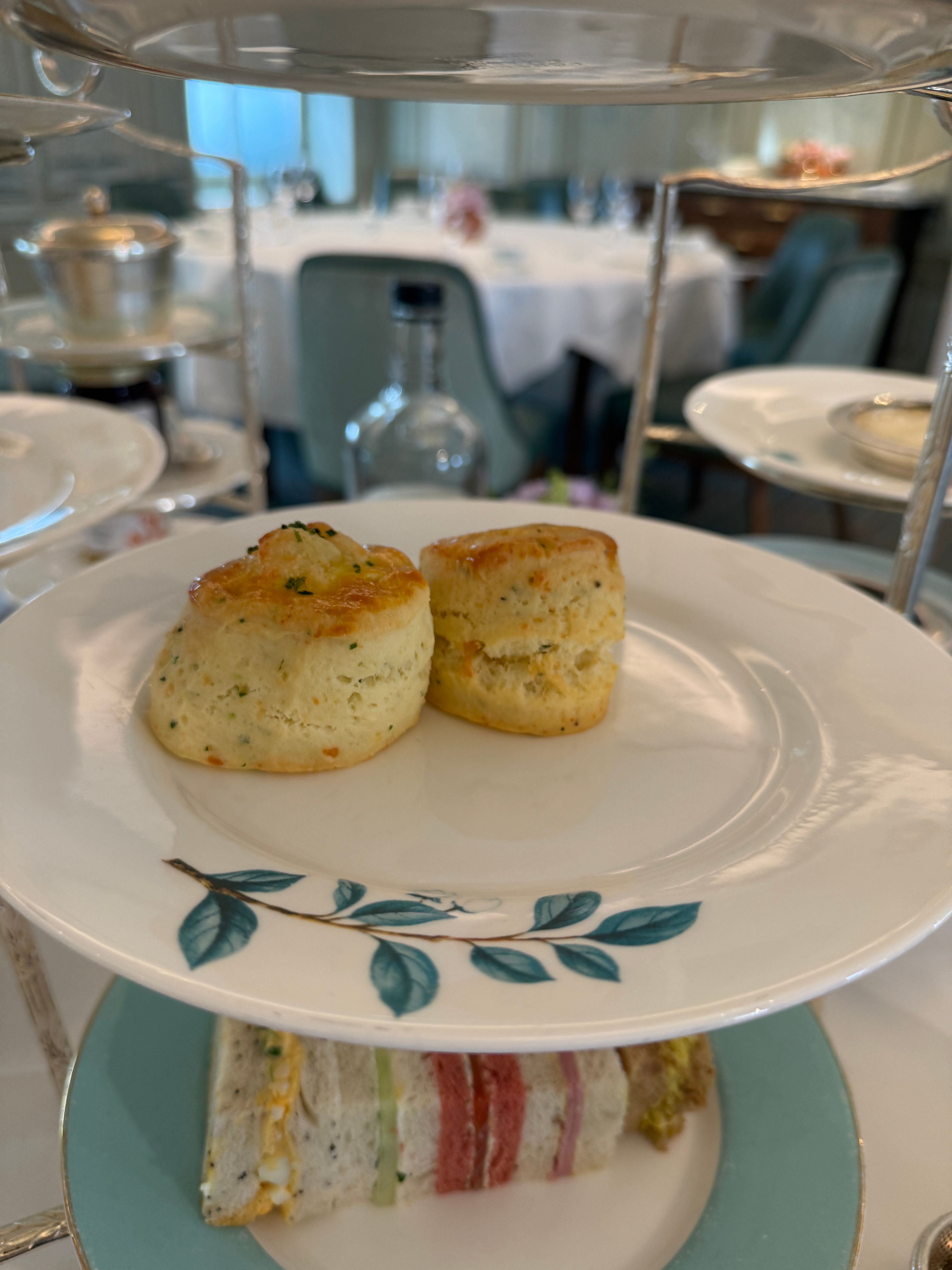 The image showcases a beautiful afternoon tea setting, with a focus on two perfectly baked scones resting on a delicate plate adorned with a leaf motif. The scones, golden and slightly flaky, evoke a sense of warmth and comfort, suggesting a delightful treat awaiting enjoyment. Below them, a tier of colorful sandwiches—each with distinct fillings—adds a playful aspect to the presentation.

In the background, a softly lit dining area hints at an elegant tea room, perhaps a luxury hotel or a quaint café, where patrons might indulge in this classic tradition. The muted colors and soft furnishings create an inviting atmosphere, ideal for leisurely conversations. 

Humorously, the scones seem to be perched like royalty on their porcelain throne, holding court over the sandwiches below, perhaps plotting a delicious takeover of the tea scene!