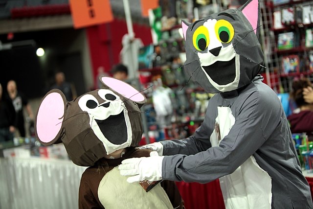 This vibrant image captures two individuals joyfully cosplaying as the iconic cartoon duo, Tom and Jerry, likely at a bustling fan convention. The person in the Tom costume, featuring an exaggerated, wide-open grin and large green eyes, gently places their gloved hands on the shoulders of the smaller Jerry, whose costume also sports a cheerful, open-mouthed expression and prominent pink ears. Their friendly interaction is a humorous subversion of their usual cat-and-mouse antics. The blurred background reveals a typical convention setting with merchandise stalls and other attendees, providing a fitting context for these beloved characters to come to life in such an endearing and playful manner.
